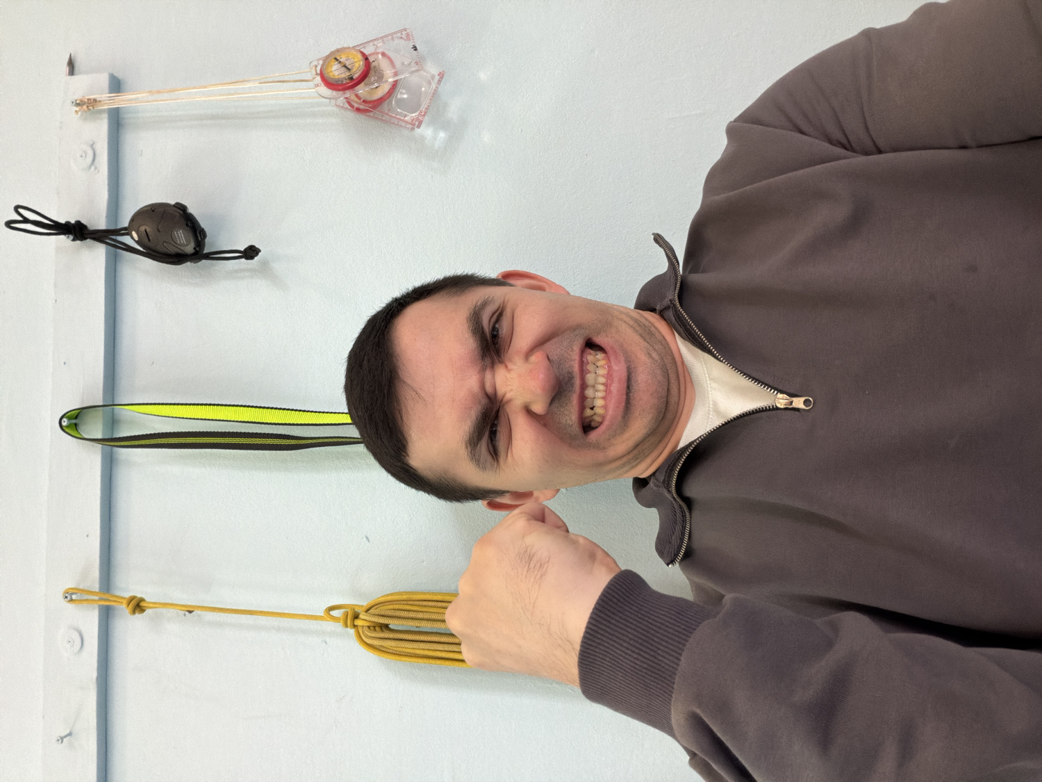 The image features an adult man posing indoors against a plain light blue wall. He has short, dark hair and is wearing a dark brown quarter-zip pullover over a white t-shirt. The man exhibits a forceful expression, with a wide grimace, bared teeth, and his right hand clenched into a fist, conveying intensity or a humorous show of strength.
Behind him, several items are visibly hanging vertically on the wall from small hooks or fasteners. These objects, from top to bottom (if the image were oriented upright), include a clear-based orienteering compass, a small black electronic device (possibly a headlamp or a compact GPS unit) attached to a black lanyard, a yellow and black webbing sling or runner, and a neatly coiled yellow rope or cord, secured at the top with a knot.
The collection of gear suggests the man has an interest in outdoor activities such as orienteering, hiking, or climbing. The setting is an indoor space, with no specific time of day or weather discernible. No visible text is present in the image. The location context provided is Izhevsk, Russia.