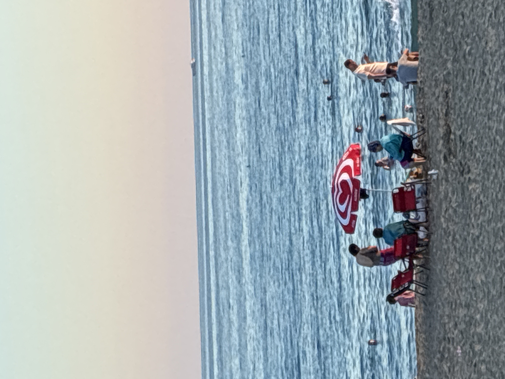 This is a medium shot image taken at a beach in Alanya, Turkey, likely during the late afternoon or early evening. The scene depicts a sandy beach with people relaxing and swimming in the sea.
In the foreground, the sandy beach occupies the bottom of the frame. A red beach umbrella with white heart designs is set up. Several red beach chairs are positioned around the umbrella.
Several individuals are present. To the left of the umbrella, a person in a light-colored shirt and patterned shorts stands near two red chairs. Another person, wearing a blue top and shorts, is seated in one of the red chairs under the umbrella. A third person, in a pink shirt, is partially visible near the water's edge to the far left, behind the umbrella.
Further back, a man in a light-colored short-sleeved shirt and dark shorts stands near the water, looking at his phone. A person in a grey t-shirt is seated on the sand behind him, facing away from the camera.
The sea is a prominent feature, taking up the middle and background of the image. It is calm with gentle waves near the shore. Several people are visible swimming and wading in the water. In the far distance, a small boat or ship can be seen on the horizon.
The sky above transitions from a pale blue at the top to a soft yellow and pinkish hue near the horizon, suggesting the time of day. The overall atmosphere is one of a relaxed beach scene.