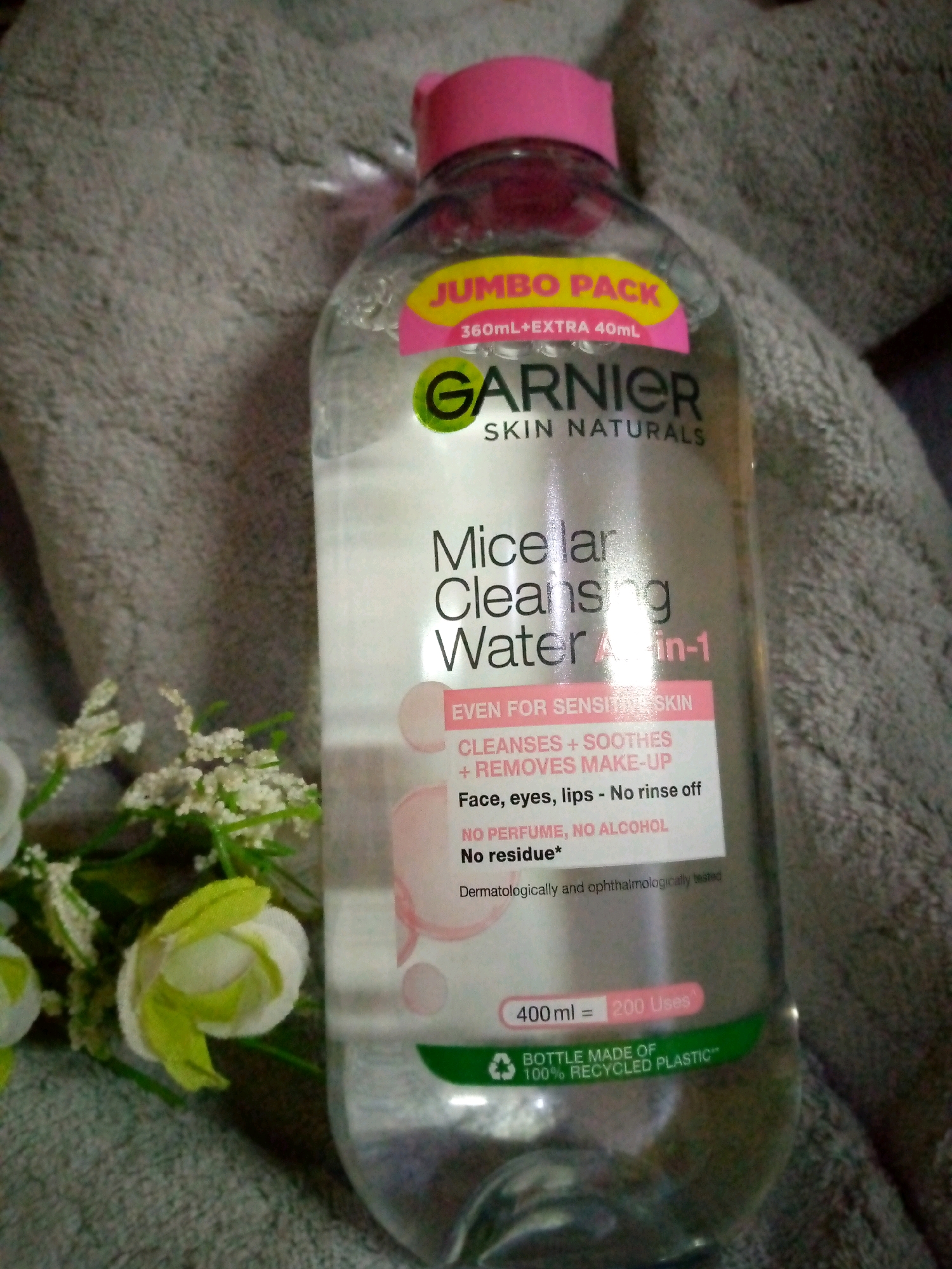 A bottle of Garnier Micellar Cleansing Water is placed on a gray, textured surface, possibly a towel or blanket, with a blurred floral arrangement to the left. The bottle has a pink cap and a prominent yellow "JUMBO PACK" label. Text on the bottle indicates it is "SKIN NATURALS," "Micellar Cleansing Water All-in-1," and is "EVEN FOR SENSITIVE SKIN." It claims to "CLEANSES + SOOTHES + REMOVES MAKE-UP" for "Face, eyes, lips - No rinse off," with "NO PERFUME, NO ALCOHOL" and "No residue*." The bottle contains 400 ml and is advertised as providing "200 Uses." A green label at the bottom states, "BOTTLE MADE OF 100% RECYCLED PLASTIC**." The image was captured in Sumberwono, Indonesia.