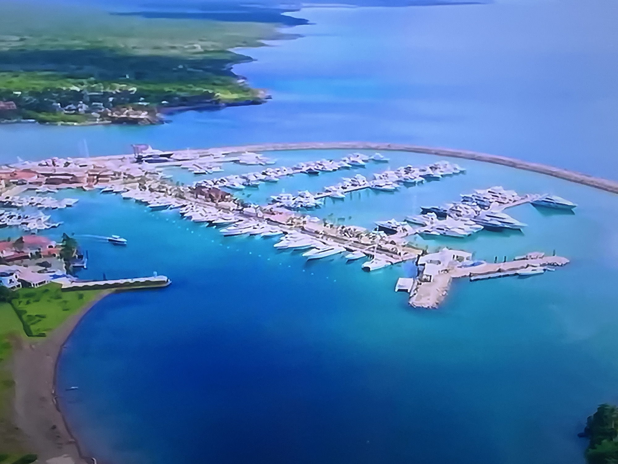This is an aerial, high-angle drone shot of a marina filled with numerous yachts docked along a curved pier. The water is a clear turquoise, and the boats are white with some visible superstructures. Beyond the marina, there's a coastline with green vegetation and buildings. The image also shows open blue water extending to the horizon. The location is a marina in an unspecified city.