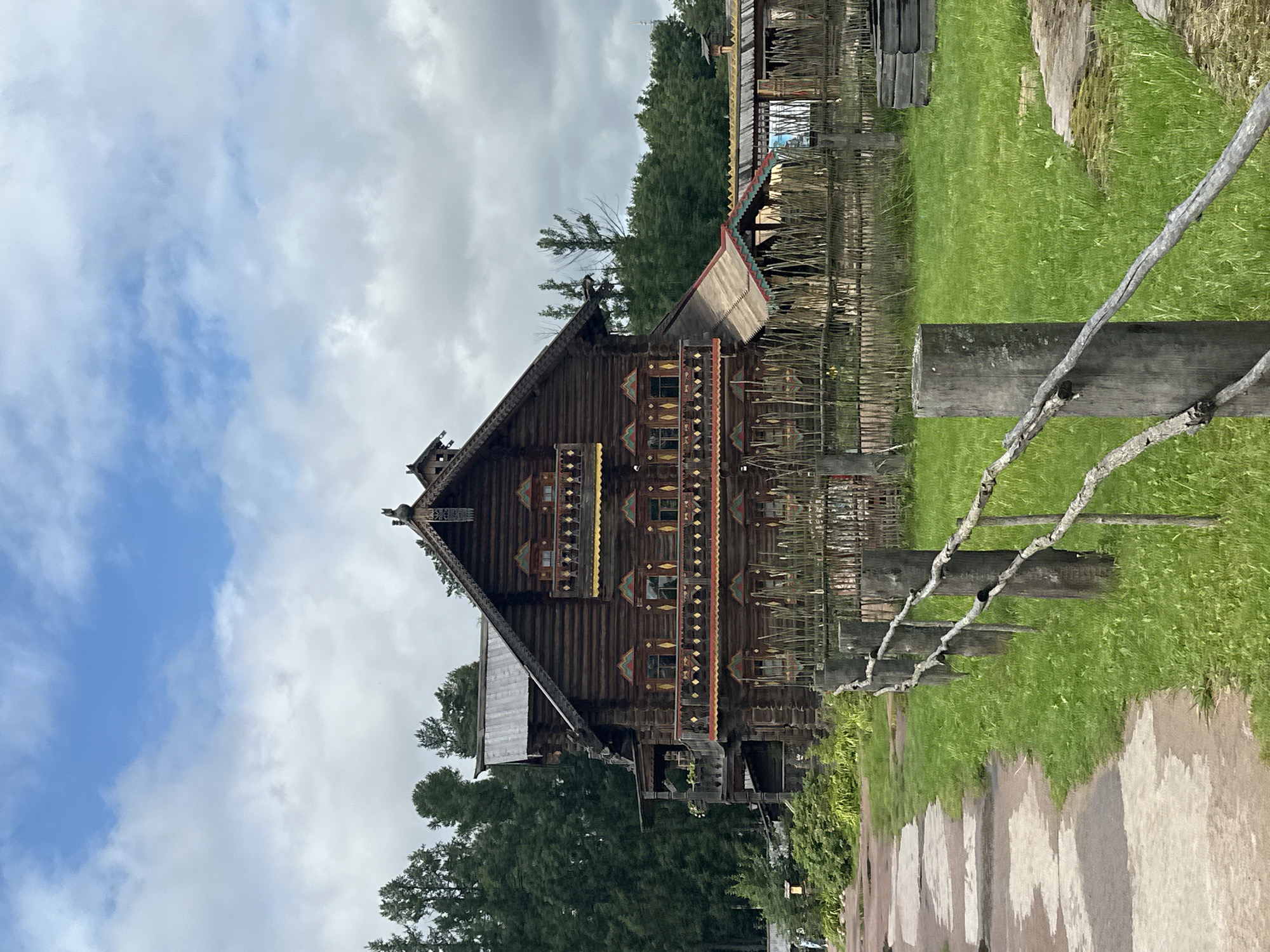 In Nevskii Parkleskhoz, Russia, a traditional wooden house stands prominently. The house is constructed of dark, horizontal logs and features ornate balconies with colorful trim in red, yellow, and green. Windows are decorated with similar colorful, triangular motifs. A steep, dark roof is topped with decorative wooden elements.
In front of the house, a rustic wooden fence made of posts and branches runs diagonally across the scene. Beyond the fence, a vibrant green lawn stretches out. A paved pathway, appearing wet in patches, leads into the frame from the left, curving around the grassy area.
Behind the house, a dense forest of dark green trees forms a backdrop. The sky above is a mix of blue and overcast white clouds. The overall atmosphere suggests a historical or cultural heritage site. There are no people visible in the image, and no specific actions or interactions are depicted.