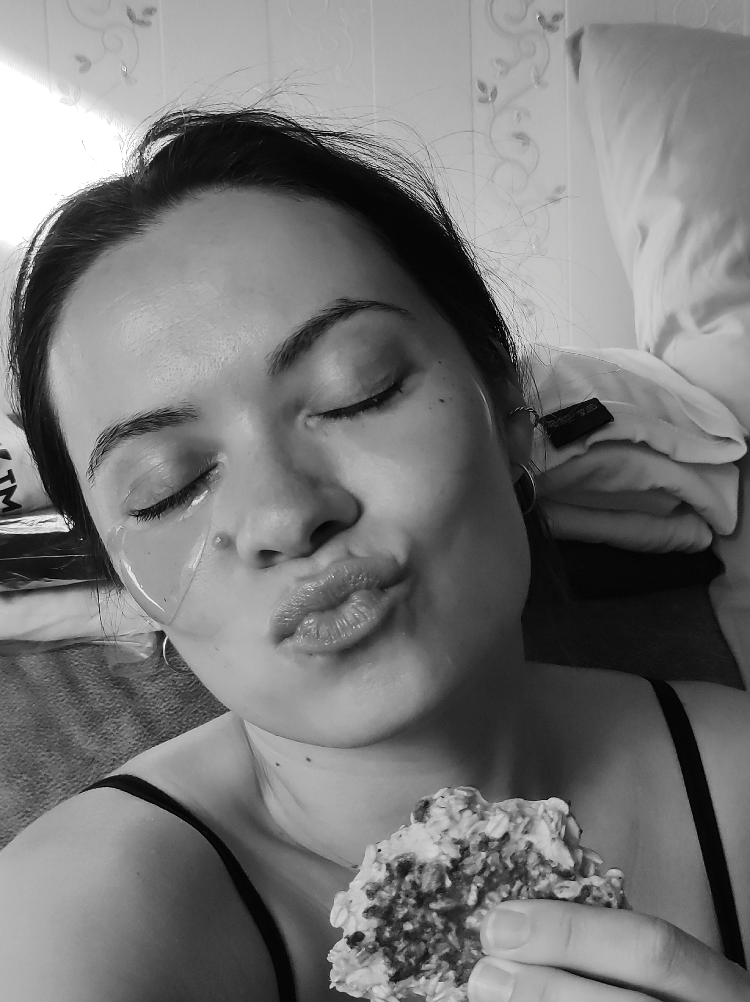 A woman is pictured from the chest up in a black and white selfie. She has eye masks on and is holding a food item in her right hand while pursing her lips in a kiss. The background appears to be a bedroom interior, with pillows visible behind her. The location is in the city of Chaikovsky, Russia.