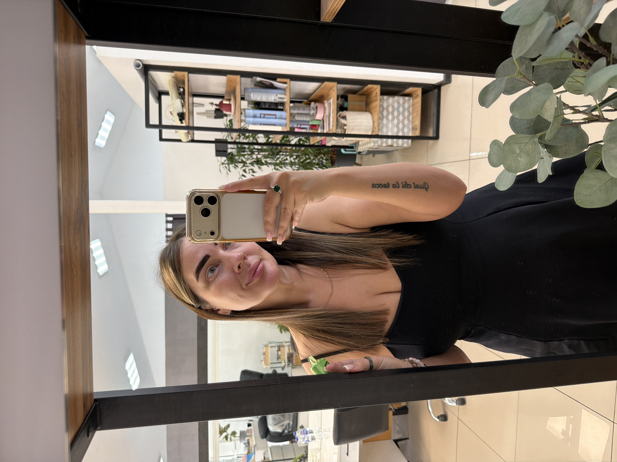 A woman is taking a selfie in a mirror. She is wearing a black sleeveless top and has a necklace on. She is also wearing a ring on her right hand and a bracelet. Her left arm has a tattoo. Her hair is brown and she has blue eyes. She is smiling at the camera. In the background, there are shelves with various items on them, including books, bottles, and boxes. There is also a plant in the foreground. The scene appears to be indoors, possibly in a salon or a room with shelves. The lighting suggests it is daytime. There is some text visible on the woman's arm, but it is not legible.