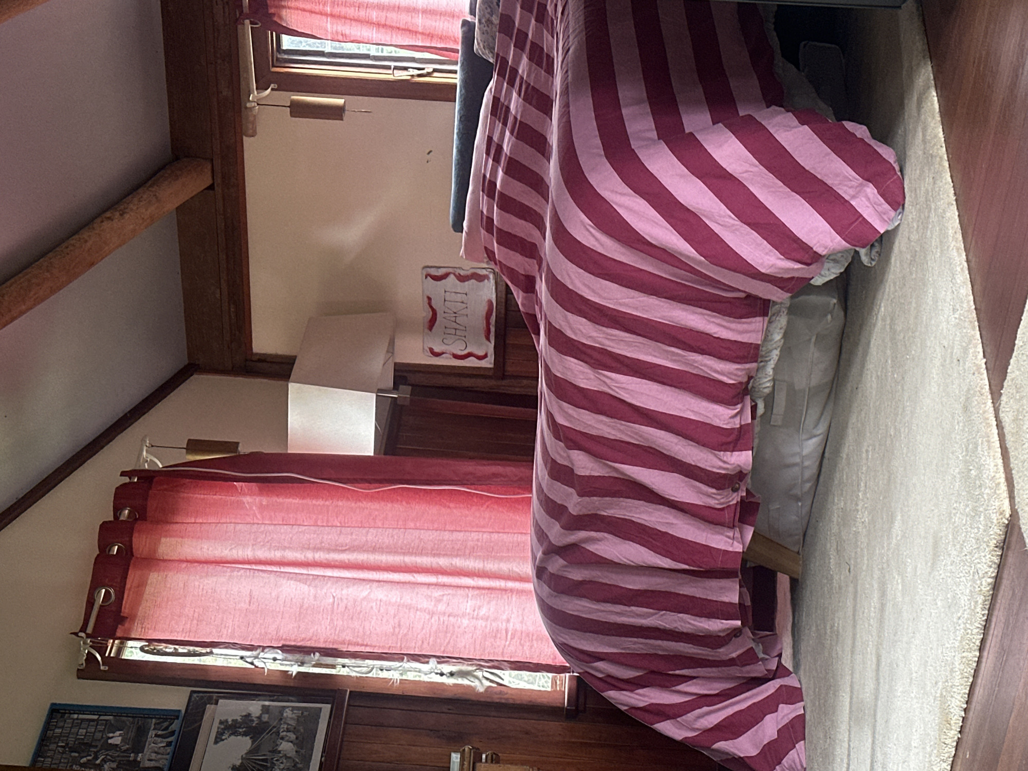 The image depicts a bedroom interior, likely in a cabin or rustic dwelling, given the exposed wooden beams and paneling. The focal point is a bed covered with a duvet featuring bold pink and dark red stripes. The bed is adorned with several pillows, one of which is a dark teal color. To the left of the bed, a window is partially covered by red curtains, and above it hangs a framed black and white photograph. To the right of the bed, a wall features a small, decorative plaque with the word "SHAKTI" written in red calligraphy. A lamp with a white shade is positioned on a bedside table or shelf. The flooring is a combination of a light-colored rug and dark wood. The environment appears to be well-lit, suggesting daylight. There are no people visible in the image, and no activities are taking place. The scene is tidy and appears to be a domestic setting. There is no information provided to suggest this location is in Thora, Australia.