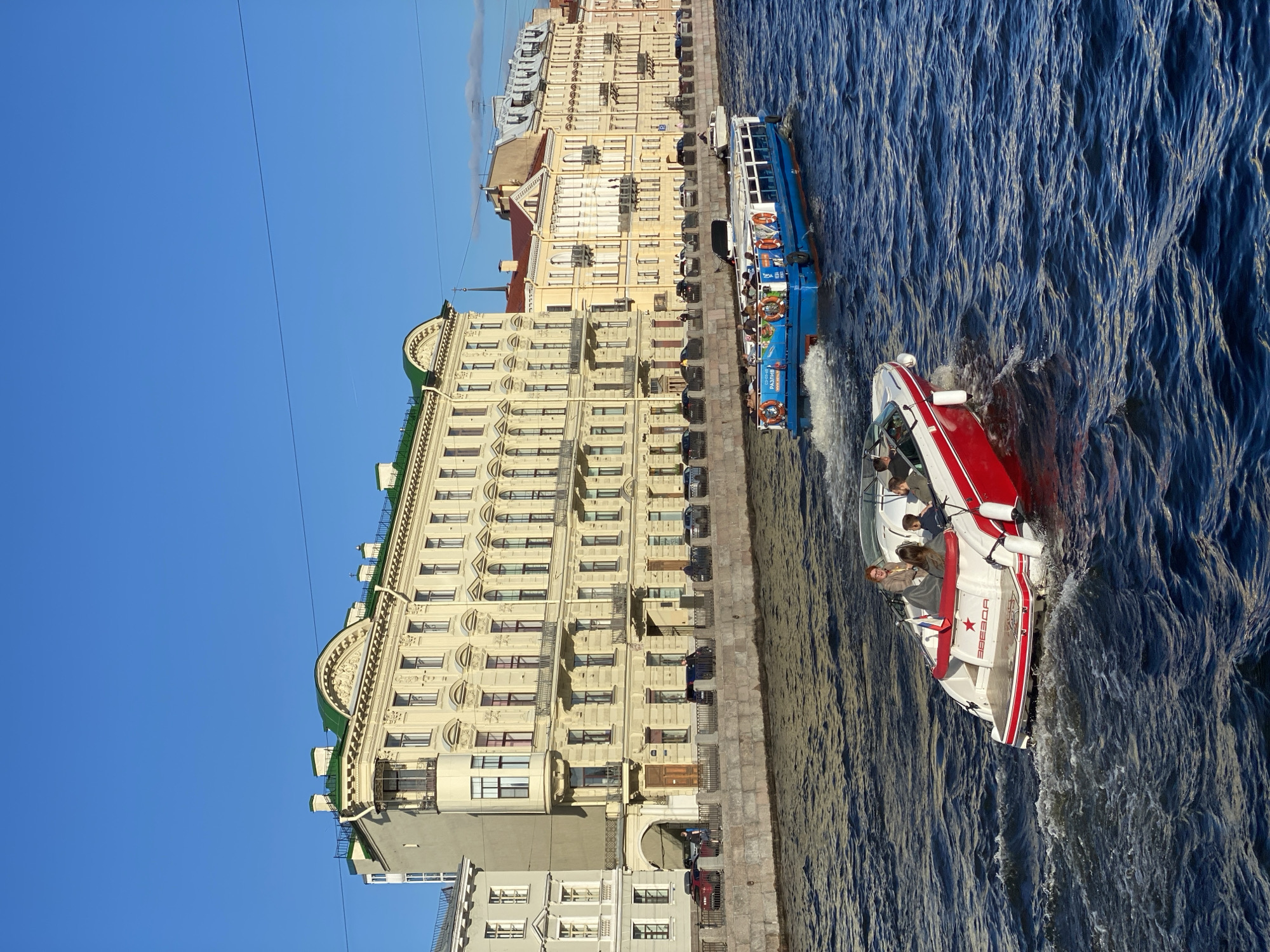 In Saint Petersburg, Russia, the image captures a sunny day along a canal. The water is a deep blue, rippling with the movement of two boats. In the foreground, a white and red speed boat carries four passengers, its wake churning the water. Behind it, a larger, blue and white tour boat, emblazoned with "РАЗЛИВ" and life preservers, is also navigating the canal.
The canal is bordered by a stone embankment, behind which a series of cream-colored buildings line the street. These buildings exhibit classic St. Petersburg architecture with ornate facades, arched windows, and green-tiled roofs. Several cars are parked along the street, and a few pedestrians can be seen walking on the embankment. The sky is a clear, vibrant blue, indicating good weather. The scene evokes a sense of urban life and leisure in the historic city.