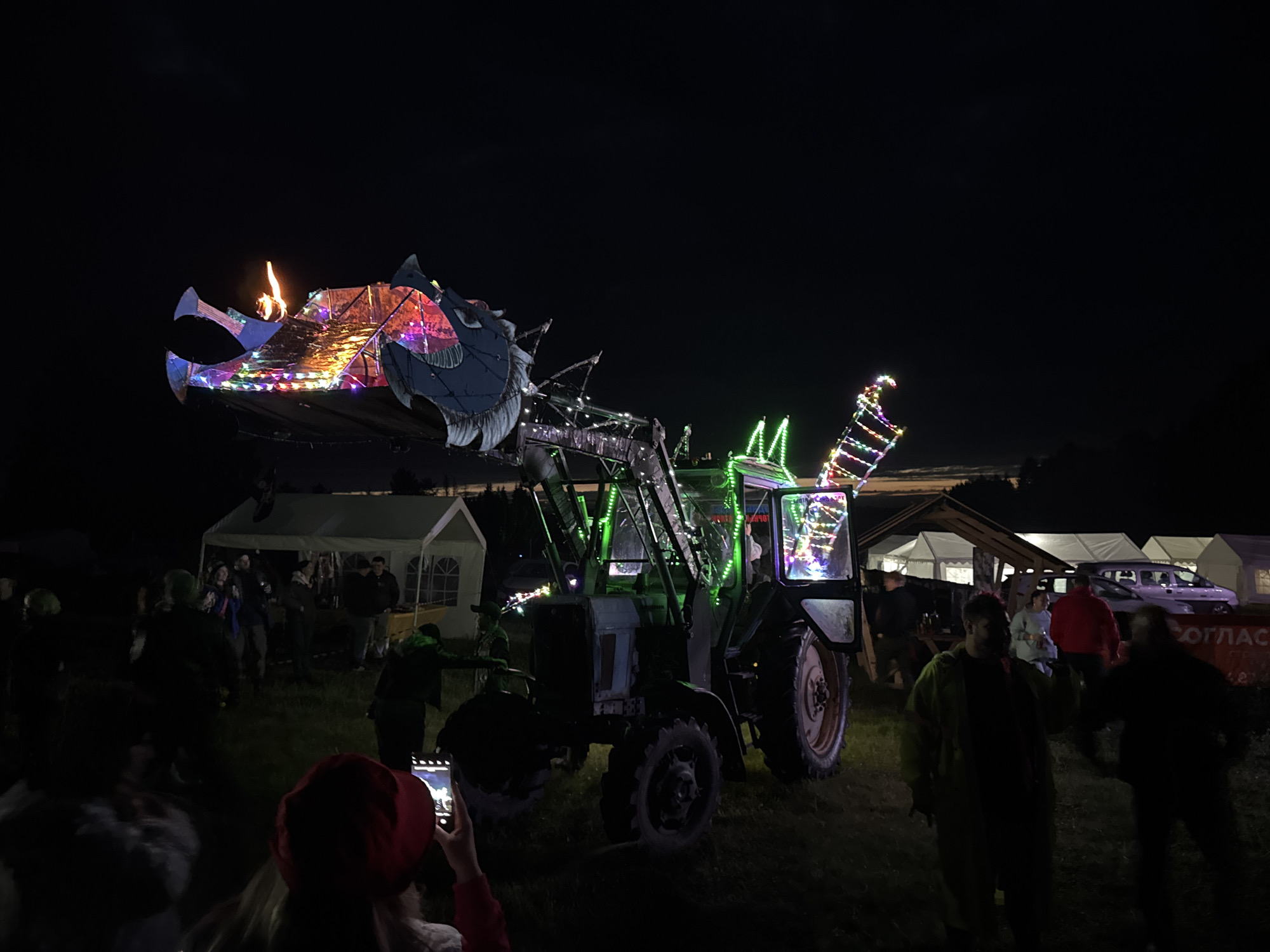 At night, in the city of Большая Кивара, Russia, an outdoor event is captured on a grassy field. The central subject is an old-model tractor, heavily modified and illuminated, positioned in the mid-ground. Its front loader is equipped with a large, elaborate structure resembling a dragon's head, adorned with multi-colored string lights (red, green, blue, yellow, pink, purple) and featuring a visible flame emanating from its top. The tractor's cabin is illuminated with green lights, and additional multi-colored string lights cover its frame and a rear extension. The vehicle has large, dark tires.
Multiple individuals are present on the field around the tractor and in the background. In the lower-left foreground, a person wearing a red head covering is seen from behind, holding a smartphone and appearing to photograph or record the scene. Other individuals are partially obscured by shadows and distance, standing and mingling. One person in the mid-ground, to the right of the tractor, is wearing a yellow jacket.
In the background, several white canopy tents are visible, illuminated from within or by external sources. To the far right, a white van or SUV is parked near one of the tents. A red sign with white Cyrillic text, partially legible as "ОГЛАС," is visible on the far right at ground level. The sky is dark and clear, indicating nighttime conditions.
