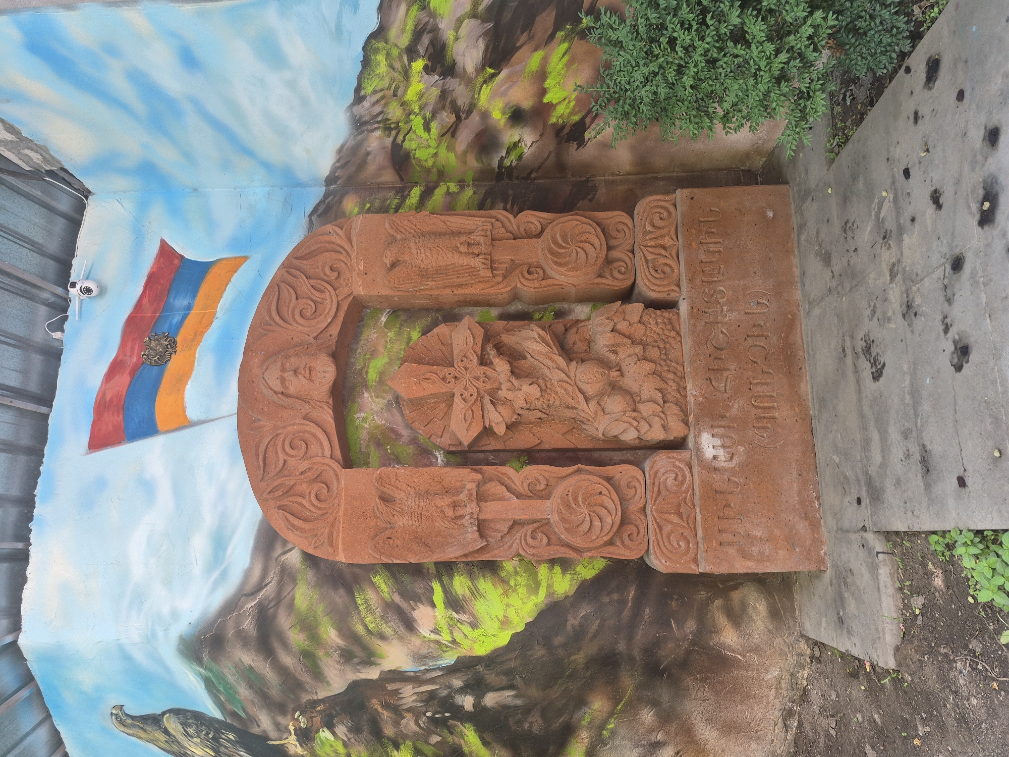 The image captures an outdoor scene with a stone monument in front of a mural. The monument is reddish-brown and features intricate carvings, including a central cross with foliage and two side panels depicting eagles. Above the monument, a mural on the wall depicts a landscape with mountains, an eagle, and the Armenian flag waving. A security camera is visible mounted on the wall above the mural. The ground in front of the monument is paved with concrete slabs. To the right of the monument, a green bush is present. The overall setting appears to be an exterior wall, possibly in an urban or semi-urban environment, given the presence of the mural and the security camera. The inscription on the monument is in Armenian. The scene is well-lit, suggesting it was taken during the day. This monument is likely a khachkar, a carved Armenian cross-stone. The mural and the khachkar are situated on a wall in Yerevan, Armenia.