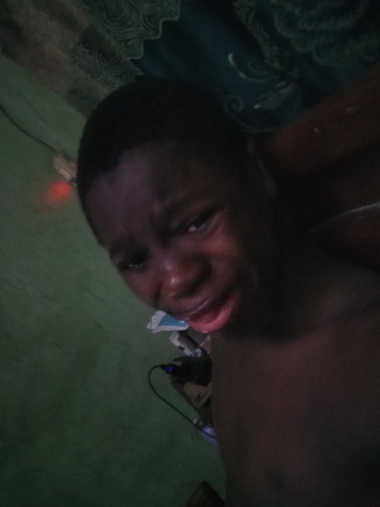 A close-up, low-angle photograph shows a young, dark-skinned child with a distressed facial expression. The child appears to be crying. The child's upper torso is visible, bare-chested. Behind the child is a green wall with what appears to be an electrical outlet and wiring. Above and behind the child's head, there's a dark, patterned curtain or fabric. The location of the scene is not discernible in this image. The city of Shangisha, Nigeria, cannot be verified from this photograph alone.