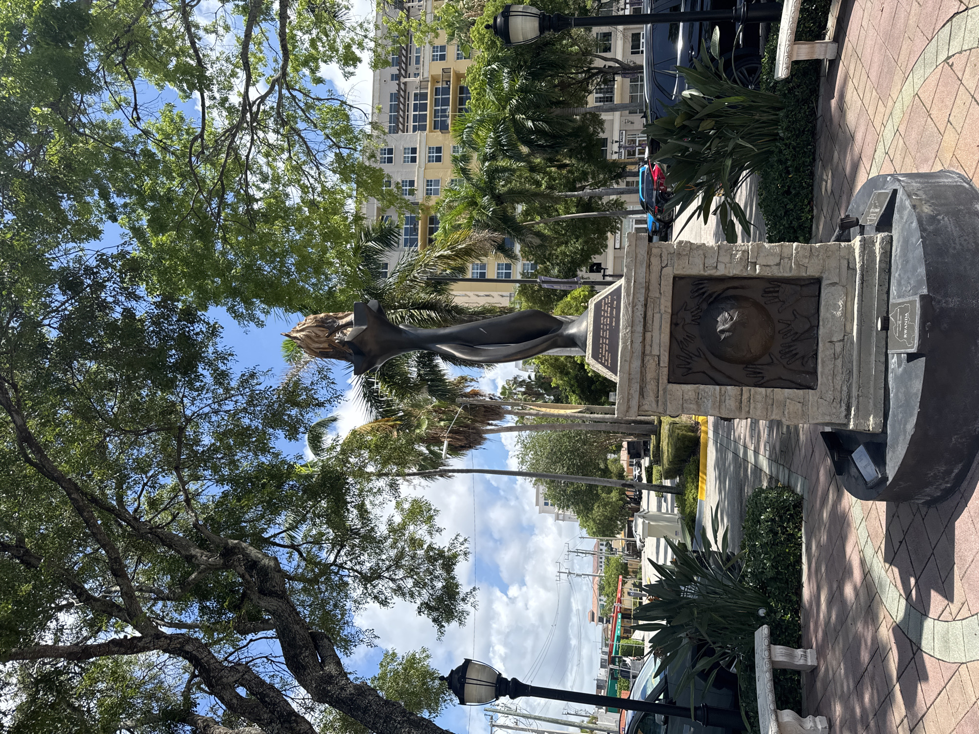 This detailed image depicts a sunlit urban park scene in Boca Raton, United States of America. At the center, a tall, abstract bronze statue rests upon a multi-tiered stone pedestal. The statue is stylized, depicting an upward-reaching form culminating in a flame-like element. The main stone pedestal features a bronze relief plaque on one side, illustrating a globe encircled by several hands with a dove above, accompanied by an inscription. A separate, darker plaque on the statue's lower base is inscribed with "RWANDA."
The immediate foreground consists of a paved walkway with a geometric pattern of reddish and lighter-toned pavers. Green, dense shrubs and several broad-leafed plants, including a Bird of Paradise, surround the statue's base and line the path. Two white, ornate stone benches are situated on either side of the central pathway. Two black lampposts with traditional lantern designs stand on the left and right sides, partially obscured by foliage.
The background reveals a typical urban landscape under a bright blue sky dotted with scattered white cumulus clouds. Large, mature trees with dense green canopies dominate the upper portion of the frame, providing shade, while numerous slender palm trees are interspersed throughout the scene. Beyond the park's immediate edge, a paved street is visible, featuring multiple parked cars of various colors (including black, red, and blue). Multi-story buildings, including a prominent light-colored residential or commercial complex with numerous windows and balconies, line the street. Utility poles and power lines crisscross against the sky in the mid-distance. The overall lighting indicates a clear, sunny day, with distinct shadows cast by the statue and surrounding elements. No people are visible in the frame.