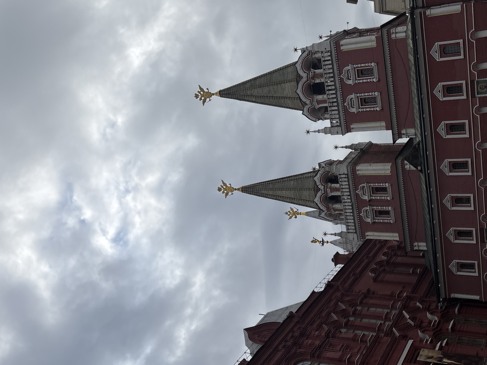 The image captures a low-angle view of the Kremlin towers in Moscow, Russia, against a cloudy sky. The towers are distinguished by their red brick walls, white decorative elements, and green, cone-shaped roofs topped with golden double-headed eagles. The architecture is ornate with various window shapes and decorative features. The sky is overcast, with layers of gray and white clouds filling most of the frame. Part of another building, also in red brick, is visible at the bottom of the image.