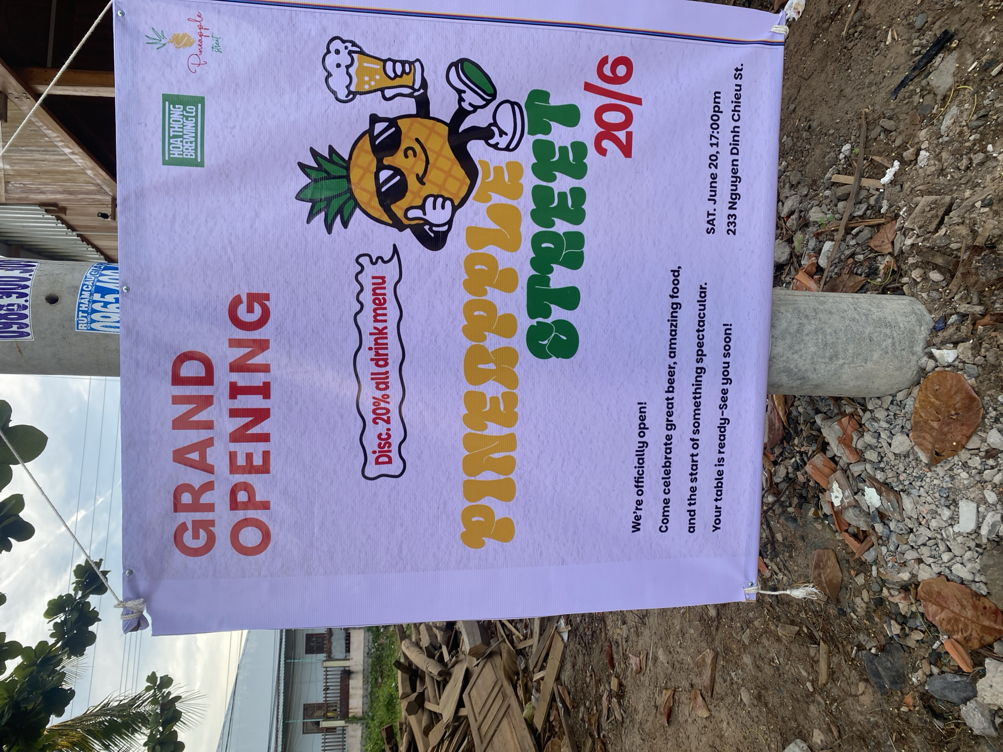 A banner advertising a grand opening is displayed on a concrete pole outdoors, near a street in Phu Thuy Ward, Vietnam. The banner is white and features red text that reads "GRAND OPENING" in large letters. Below this, it states "Disc. 20% all drink menu" in a speech bubble. The banner also includes cartoon imagery of a pineapple holding a beer and giving a thumbs-up. The text "PINEAPPLE STREET 20/6" is prominently displayed in yellow and green letters. Smaller text at the bottom announces "We're officially open! Come celebrate great beer, amazing food, and the start of something spectacular. Your table is ready—See you soon!" and provides event details: "SAT. June 20, 17:00pm 233 Nguyen Dinh Chieu St." The background shows a house, a pile of wooden debris, and a tree. The scene appears to be on a street or roadside, with the ground covered in dirt, rocks, and fallen leaves. The sky is visible in the upper portion of the image.