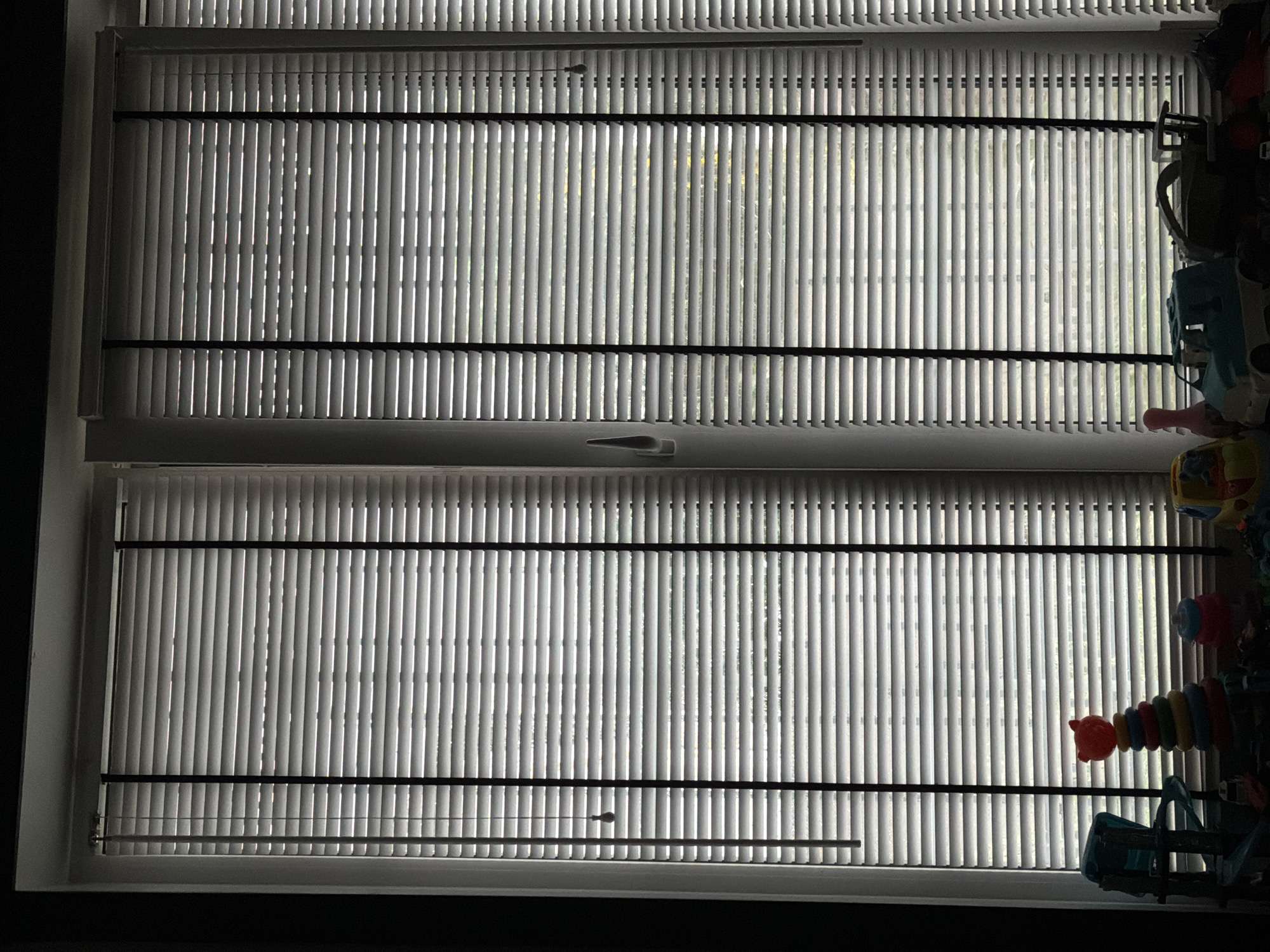 The image shows a close-up of a window with horizontal blinds partially closed, allowing some light to filter through. The blinds are white with black cords running vertically. On the windowsill below, several children's toys are visible. These include a stacking ring toy with a red bear head on top, a pink bowling pin, a blue and white toy truck, and other assorted toys that are partially obscured. The overall scene suggests a domestic indoor setting, possibly a child's room. There is no visible indication of the location being in Bila Tserkva, Ukraine, or any specific city.