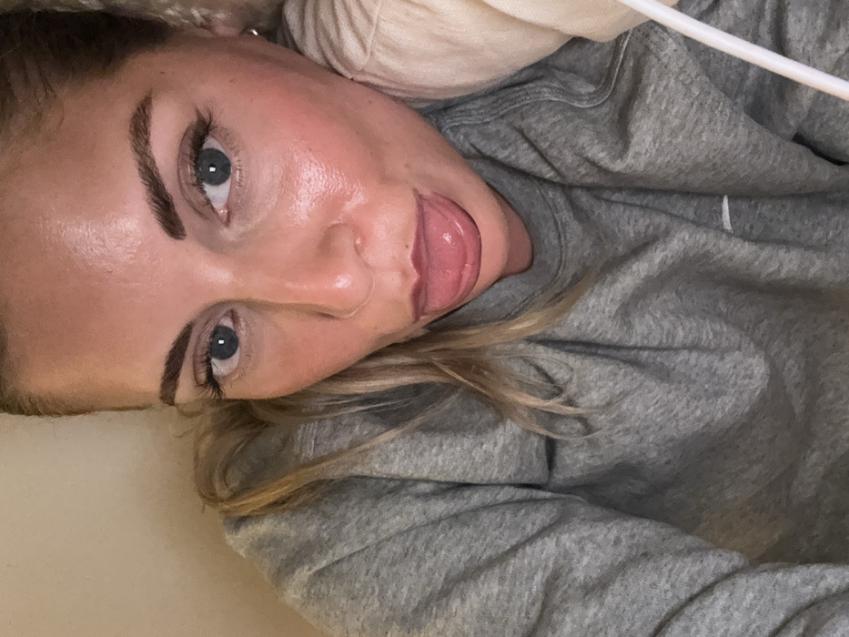 A woman with light skin, blue eyes, and defined eyebrows lies down, sticking out her tongue. She is wearing a grey hoodie. Some strands of blonde hair are visible near her shoulder. The image is taken from a close-up, slightly angled perspective, emphasizing her face and upper body. The background is plain, drawing focus to the subject.