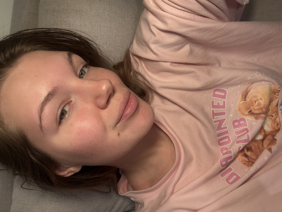 A woman lies on a gray surface, her head tilted towards the camera. She has fair skin, brown hair, and light green eyes. The woman's eyebrows are full and well-defined, and she has a small mole on her cheek. She is wearing a light pink long-sleeved shirt with a print of two bunnies and the words "Disappointed Club." The shirt is wrinkled, and the lighting in the photo is soft.