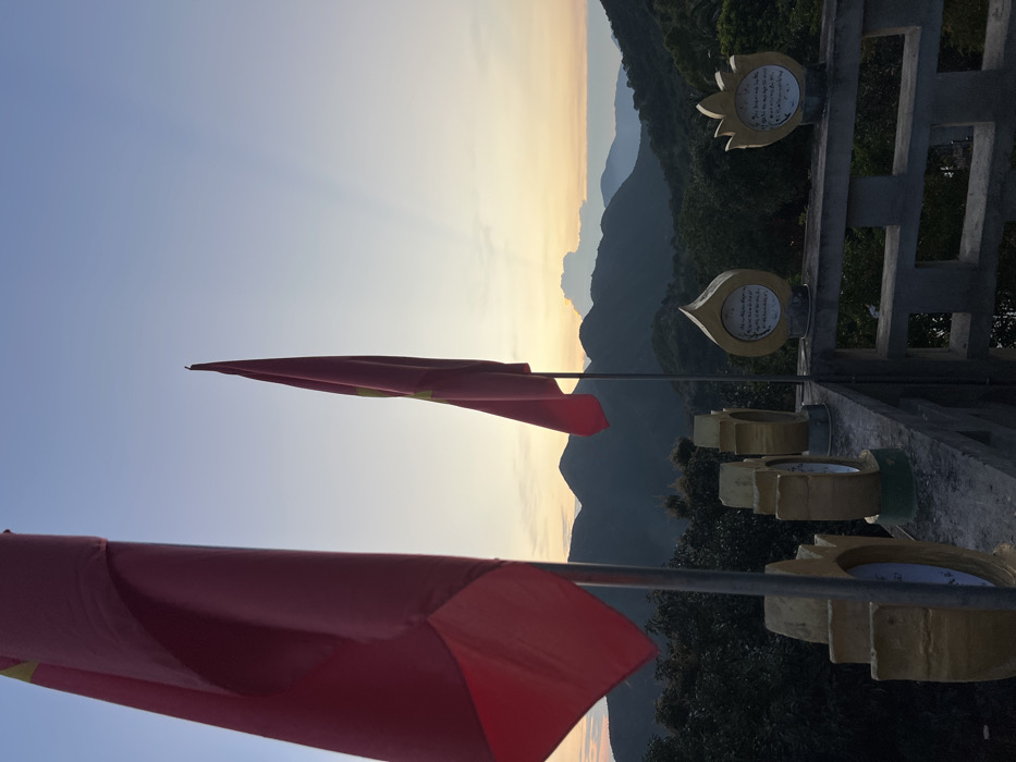 The image displays an elevated outdoor platform situated in a mountainous area of Nha Trang, Vietnam, at either dawn or dusk. Two red flags, each featuring a central yellow star, identified as the national flag of Vietnam, are prominently displayed on poles. The flag on the left occupies the far left of the frame; the second flag is positioned further right. Both flags hang partially furled. A concrete railing or balustrade forms the immediate foreground, running horizontally across the lower third of the image. Mounted on this railing are several golden-colored, stylized decorative elements. These objects vary in shape, with some resembling urns and others lotus petals; at least two contain white circular or teardrop-shaped inserts with dark script. Dense green vegetation covers the terrain below the platform and extends across the slopes of multiple layered mountain ranges visible in the background. The sky exhibits a gradient, transitioning from light blue overhead to orange and yellow near the horizon, where visible sun rays emanate from a cloud formation above the distant mountains.