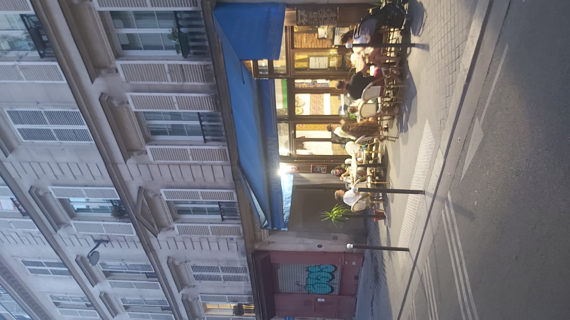 The image shows a street scene in Paris, France, at dusk. A building with multiple stories and white shutters lines the street. A restaurant or cafe with a blue awning is situated on the ground floor, with tables and chairs set up on the sidewalk. Several people are seated at the tables, engaged in conversation or eating. The street in the foreground is asphalt with white lane markings. The overall atmosphere is relaxed and casual, with the warm glow of the restaurant's interior and streetlights casting a soft light on the scene.