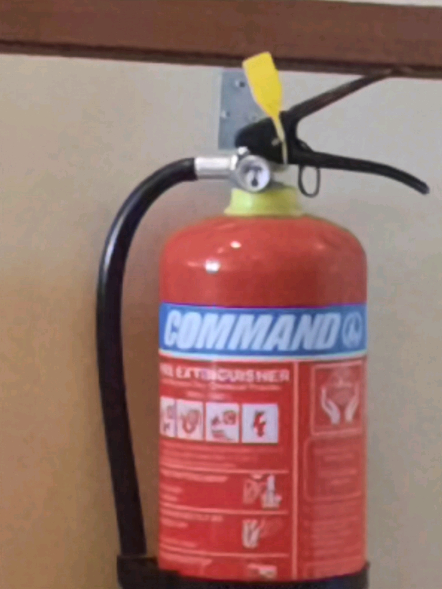 The image shows a red fire extinguisher hanging on a wall. The fire extinguisher has a blue band with white lettering that says "COMMAND" and "FIRE EXTINGUISHER". There is a yellow tag attached to the handle. The wall appears to be painted a light color. The scene is indoors.