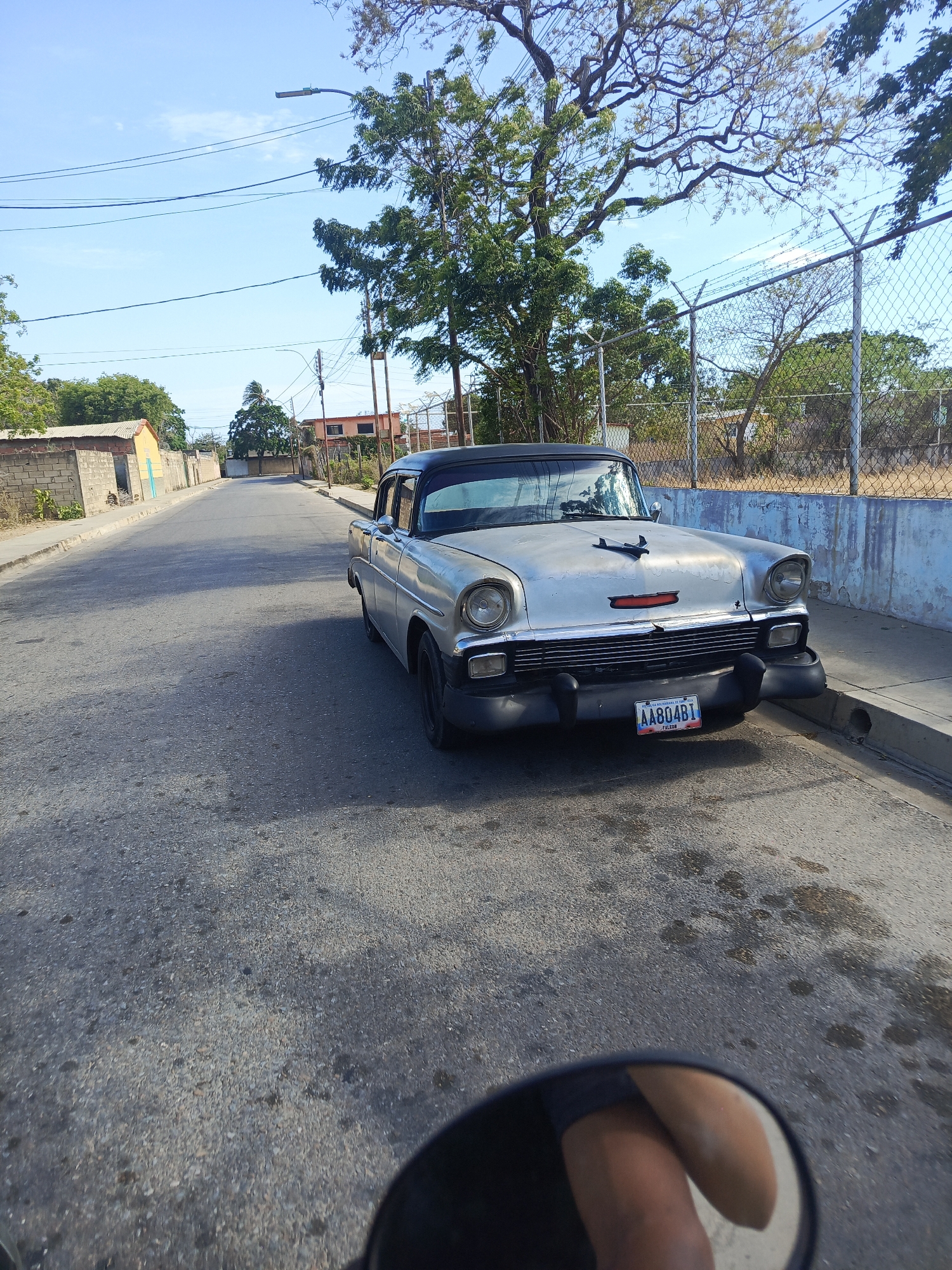 The image shows a vintage silver car with a black roof parked on the side of a street in Juan Griego, Venezuela. The car has a license plate reading "AA804BI." The street is asphalt and appears worn. A sidewalk runs alongside the street where the car is parked. A chain-link fence stands on the other side of the sidewalk. Trees and other vegetation are visible behind the fence. Some buildings are visible on the left side of the street. The sky is blue and clear. A rearview mirror, seemingly of a motorcycle or scooter, is visible in the lower part of the image, reflecting what appears to be a person's leg. The scene is outdoors and appears to be taken during the day.