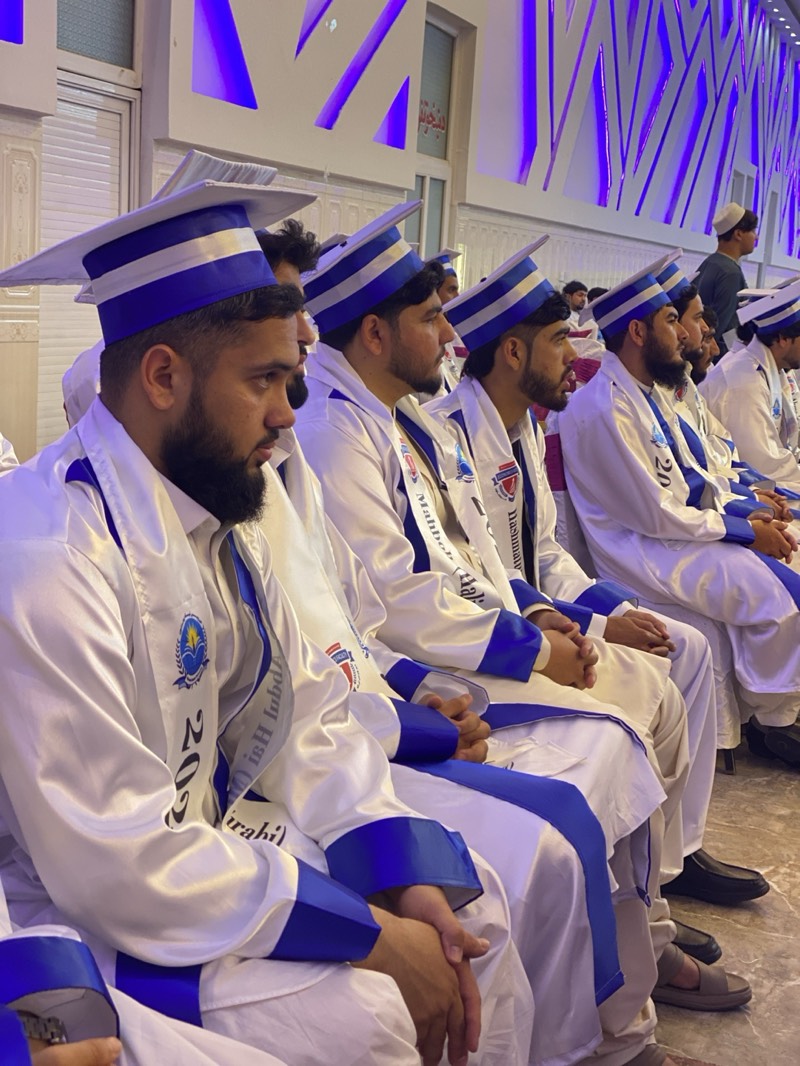 This image captures a graduation ceremony in Kandahar, Afghanistan, featuring a group of young men seated attentively in rows. The subjects are dressed in distinctive academic attire: white, satin-like gowns with blue trim on the sleeves and along the front, paired with unique blue and white striped, flat-topped graduation caps. Each individual wears a white stole with blue trim, draped over their shoulders.
Visible text on the stole of the prominent foreground subject clearly shows "2023," indicating the graduation year. Other stoles display partial Arabic or Persian script, including discernible segments such as "Mahmud," "Lubab," and "Irabi" (or similar phonetic spellings), alongside an institutional emblem or logo. The men generally have beards and dark hair, reflecting regional demographics.
The setting is an indoor hall, likely an auditorium or event space, distinguished by modern, geometric wall panels illuminated with striking blue lighting, casting a cool ambiance. The attendees are seated, looking forward, suggesting they are focused on a speaker or a central event. The overall atmosphere conveys a sense of formality, achievement, and solemnity, characteristic of a commencement ceremony. The dominant colors in the scene are white and blue.