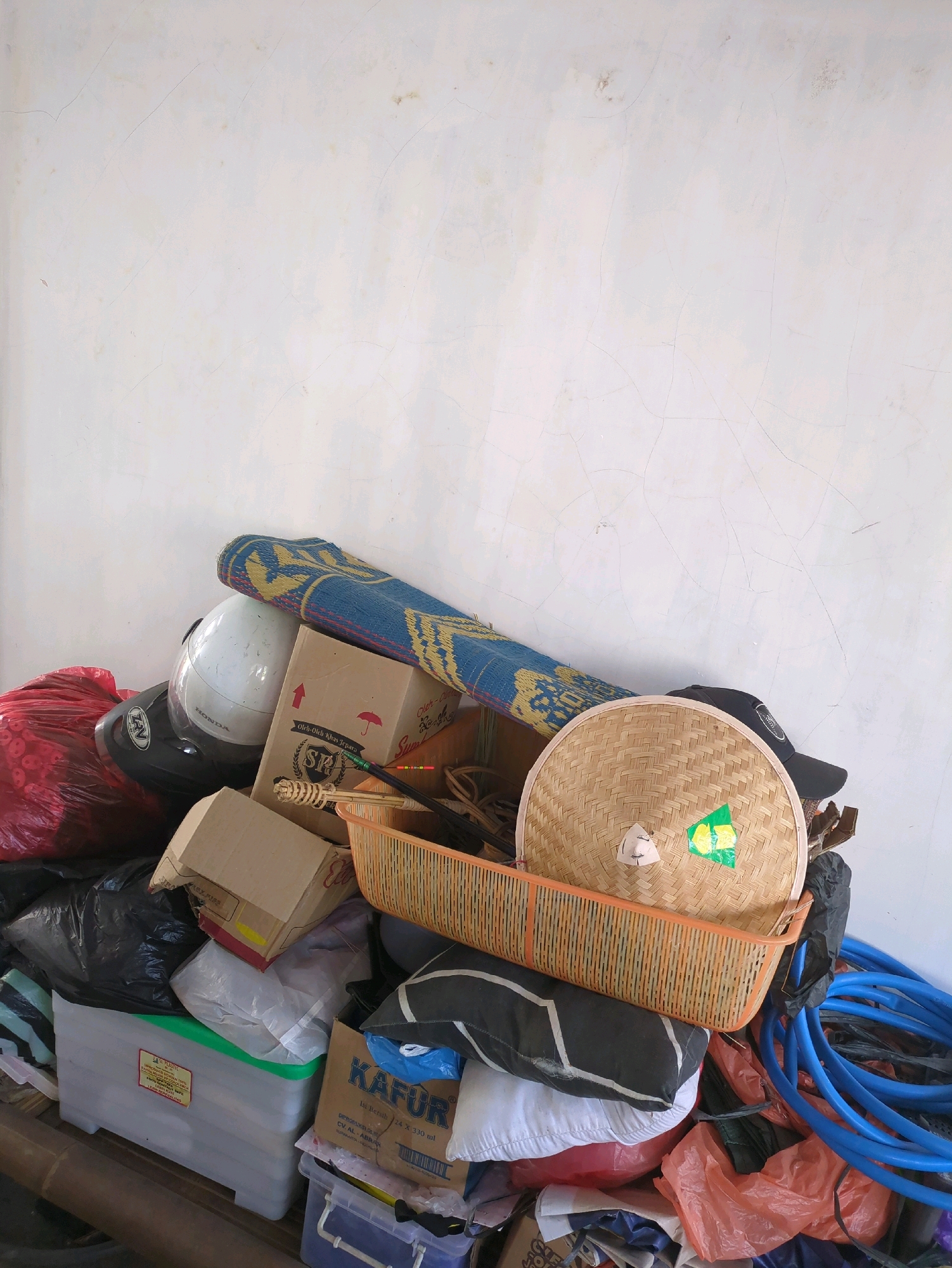 The image depicts a cluttered area against a white, cracked wall, likely in Sindon, Indonesia. A pile of various objects is stacked near the bottom right of the frame. Dominating the composition are a woven bamboo hat with green and yellow stickers on it, resting in an orange plastic basket. To the left of the hat, a white motorcycle helmet with black accents is visible, placed on top of a large red plastic bag. A blue and yellow patterned mat is draped over the helmet and basket. Several cardboard boxes, some with text and red markings, are scattered amongst the items. Black plastic trash bags, a clear plastic storage bin with a green lid, and a stack of white and black pillows are also present. To the far right, a coil of bright blue tubing is prominently displayed. The overall impression is one of disarray and accumulated possessions, typical of storage areas or spaces where items are temporarily kept. The background wall shows signs of age and weathering, with visible cracks and some faint discoloration.