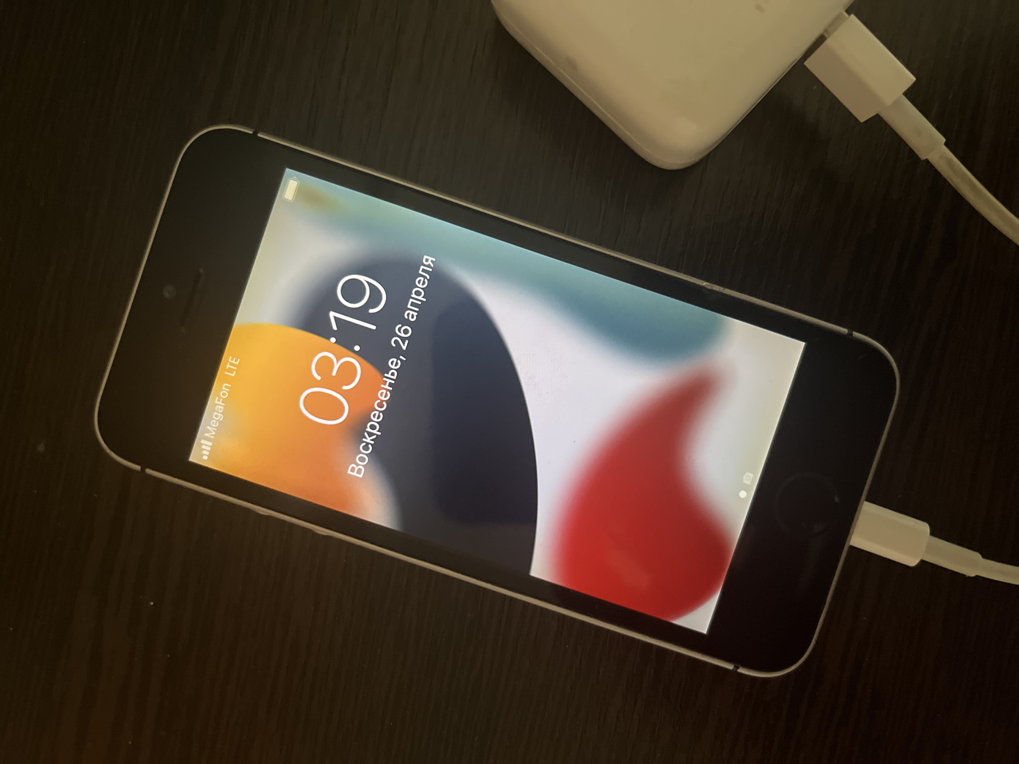 An iPhone is laying diagonally on a dark brown surface. The screen is illuminated and shows a blurred abstract wallpaper with shades of yellow, grey, white, and red. The time "03:19" and the date "Воскресенье, 26 апреля" (Sunday, April 26th) are displayed in white. The cellular network is listed as "MegaFon LTE." A white charging cable is plugged into the bottom of the iPhone, and the other end is connected to a white power adapter. The power adapter rests partially out of frame on the same dark brown surface as the iPhone. The scene is located in Chaikovsky, Russia because the cell service is listed as MegaFon, a Russian company, and the date is written in Russian.