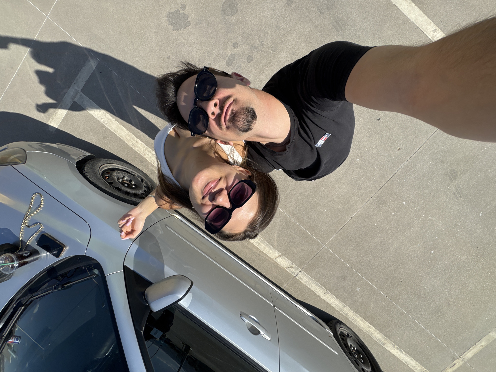 A high-angle photograph captures a couple standing near a silver car in what appears to be a parking lot. The man, wearing a black t-shirt and round sunglasses, is positioned slightly above the woman, who wears a white top and dark sunglasses. A car is located on the right side of the frame with its windshield wipers and side mirror visible. A phone, a Starbucks cup, and a gold chain are lying on the hood of the car. The shadows of the couple fall beside the car, suggesting a sunny day. The location is likely Sofia, Bulgaria, given the metadata. The parking lot is marked with white lines, indicating parking spaces.