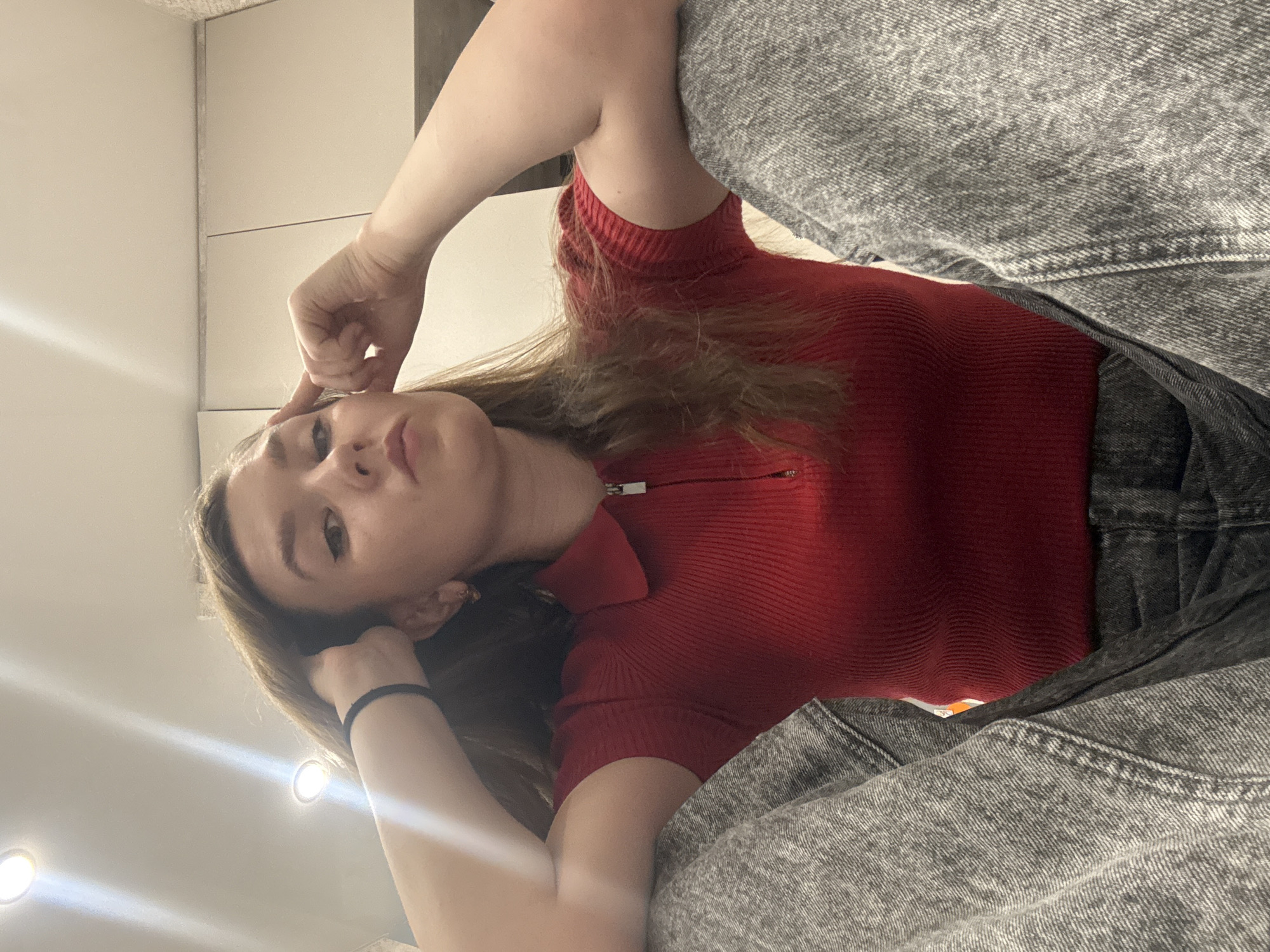 The image shows a woman with brown hair wearing a red ribbed top and gray jeans. She is in an indoor setting, possibly a room with neutral-colored walls. Her left hand is touching her head and wrist band, while her right hand is on her forehead. There is no information present that the location is in the city of Izhevsk, Russia.