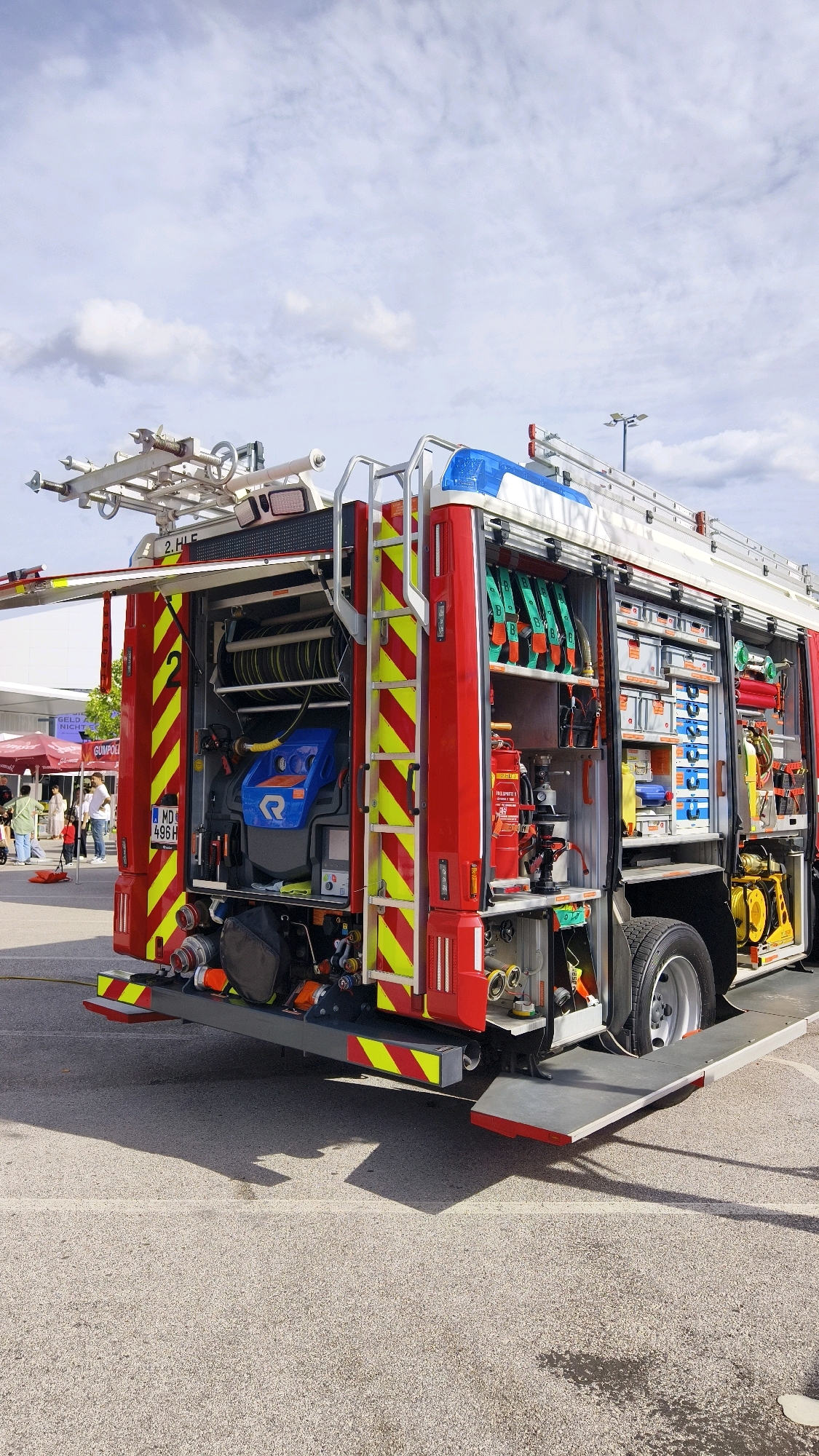 An external, eye-level, medium shot captures the rear and side of a red fire truck parked on an asphalt lot under a cloudy sky. The truck is equipped with various tools and equipment, including hoses, ladders, and storage compartments. The rear of the truck features a license plate that reads "MD 496 H." Several people are visible in the background, some standing near a building, while others are walking. The environment appears to be an outdoor public space, possibly a parking lot or a street, with some light poles visible in the distance. The overall scene is still and appears to be a static display or demonstration. This image was taken in Vösendorf, Austria.