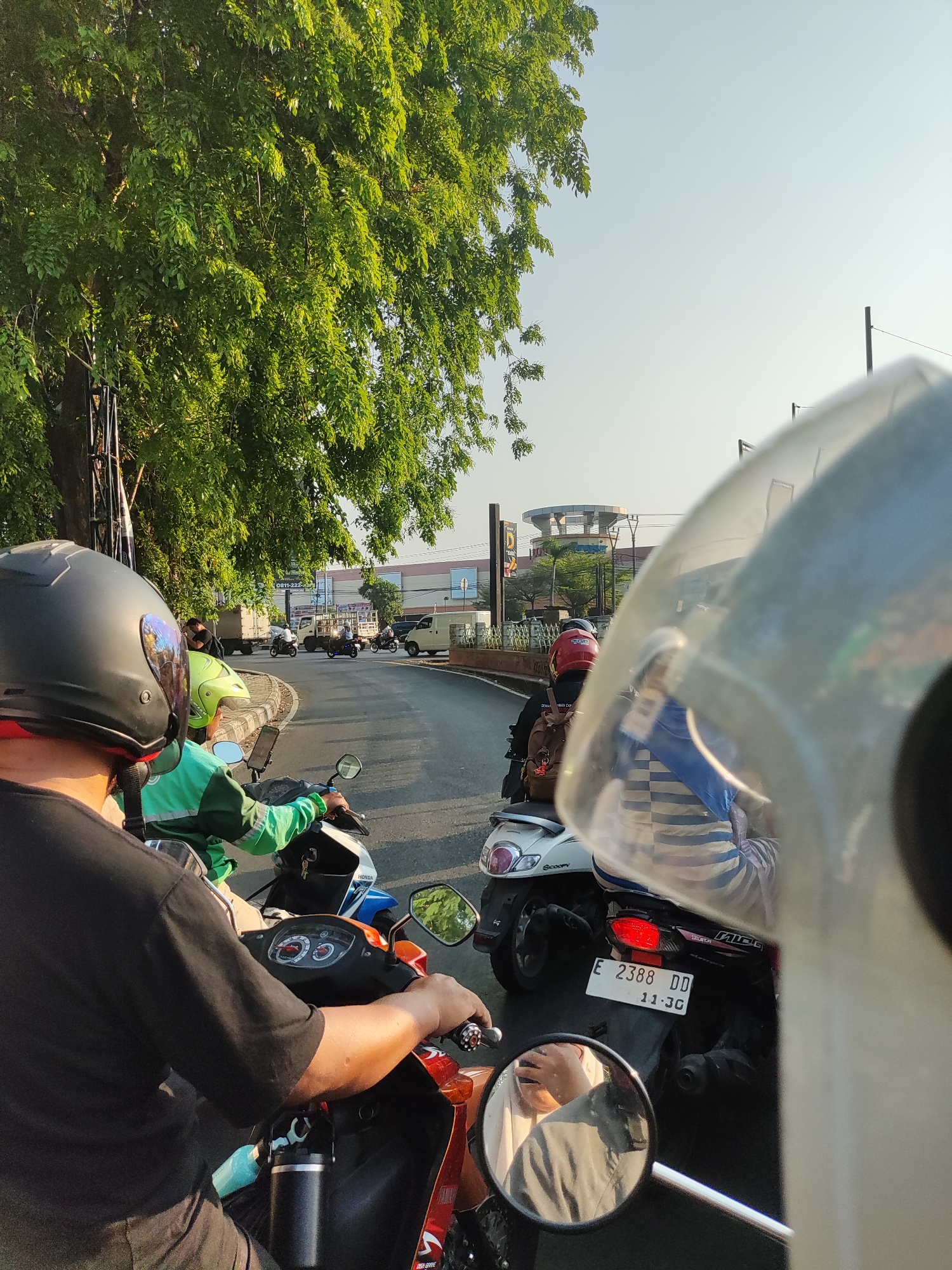 The image depicts a traffic scene in Cirebon, Indonesia. The perspective is from the rider of a motorcycle, facing forward into a street with several other motorcycles and vehicles. The rider is wearing a black helmet, and the front of a white helmet is visible in the foreground, obscuring part of the view. In front of the rider is another motorcycle with a rider wearing a bright green jacket and a lime green helmet.
The street appears to be a moderately busy road, with a gentle curve to the right. On the left side of the road, a large tree with lush green foliage casts shadows. Beyond the tree, there are some utility poles and a building with signage, one of which appears to be in Indonesian. In the distance, a large, modern building with a circular element on top is visible, likely a commercial establishment.
Several other motorcycles are in view, some carrying multiple passengers, all navigating the traffic. License plates are visible on some of the motorcycles, such as "E 2388 DD". The sky is clear and bright, suggesting it is daytime, possibly late afternoon due to the angle of the sun creating warm lighting. The overall atmosphere is that of a typical urban commute in Indonesia.