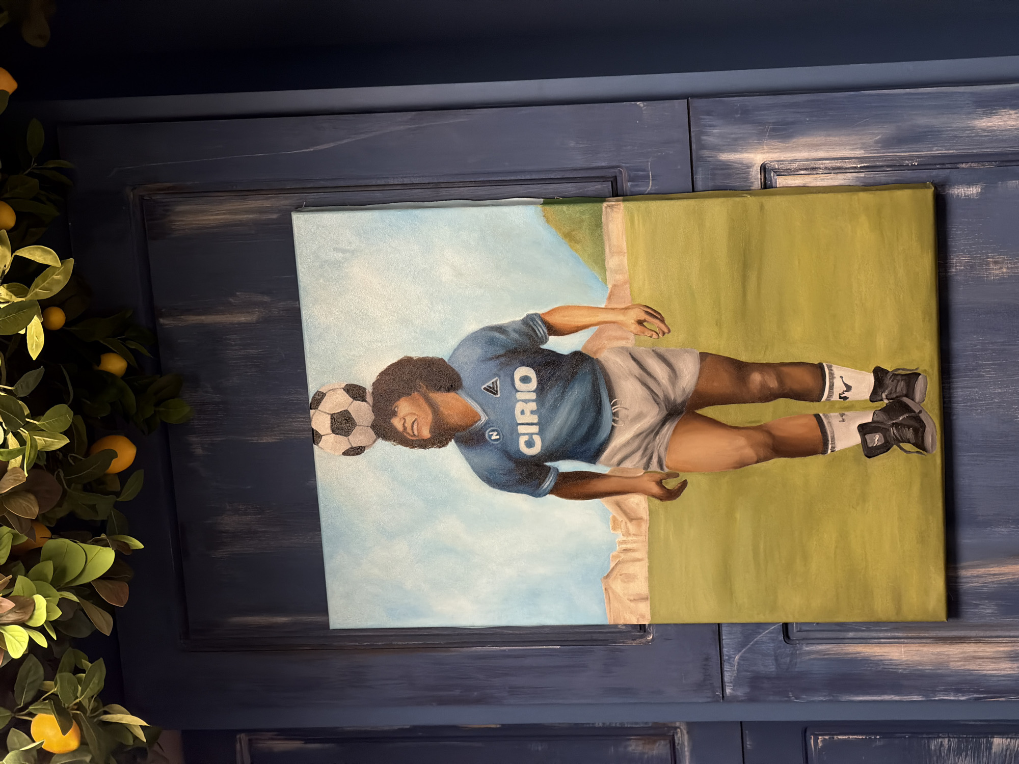 The image displays a canvas painting, rotated 90 degrees clockwise, portraying a male figure, likely a soccer player, balancing a soccer ball on his head. The figure has dark, curly hair and a smiling expression. He is wearing a light blue short-sleeved jersey with "CIRIO" written vertically in white down the front, and a small "N" logo above it, characteristic of SSC Napoli, a strong visual cue pointing to Diego Maradona. He also wears light gray shorts, white socks with black stripes, and black athletic shoes or cleats.
The painting's background features a bright light blue sky and a light green field or pitch, with a faint, light-colored horizon suggesting distant structures. The setting within the painting appears to be outdoors on a sunny day.
The canvas itself is placed against a dark blue, textured wooden surface, possibly a cabinet door or paneling, exhibiting a distressed or whitewashed effect with lighter streaks. To the left of the painting, vibrant green foliage with numerous bright yellow lemons is visible, suggesting artificial lemon branches. The lighting indicates an indoor setting for the photograph.