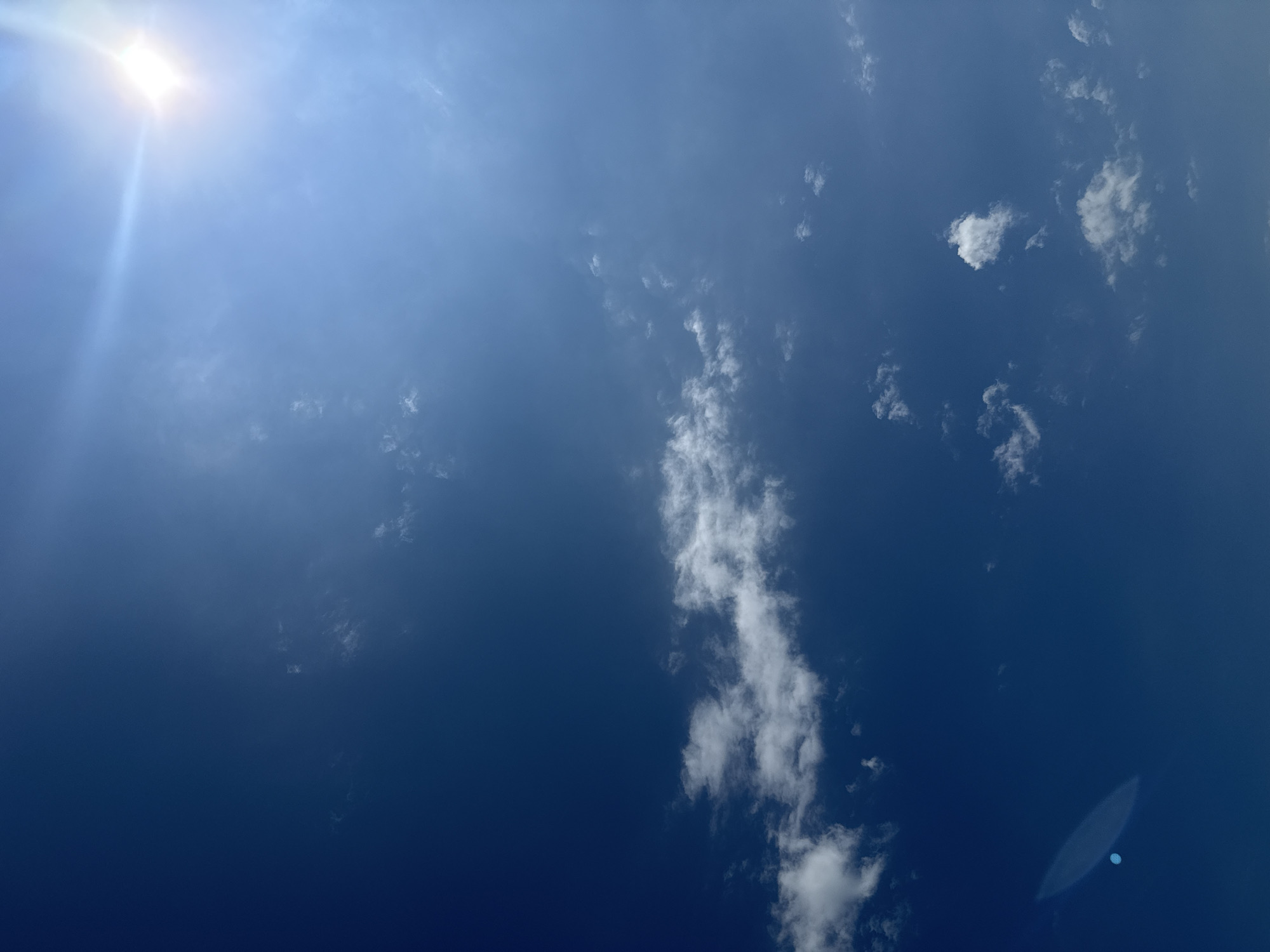 A wide shot captures a deep blue sky with scattered white clouds. The sun, a bright, lens-flare-effected orb, is positioned in the upper right quadrant, casting a bright light. In the lower left quadrant, a distinct oval-shaped glare, likely from the camera lens, is visible. The image appears to be taken outdoors during the day. Without any distinguishing landmarks, it is impossible to determine the specific location within Nags Head, United States of America.