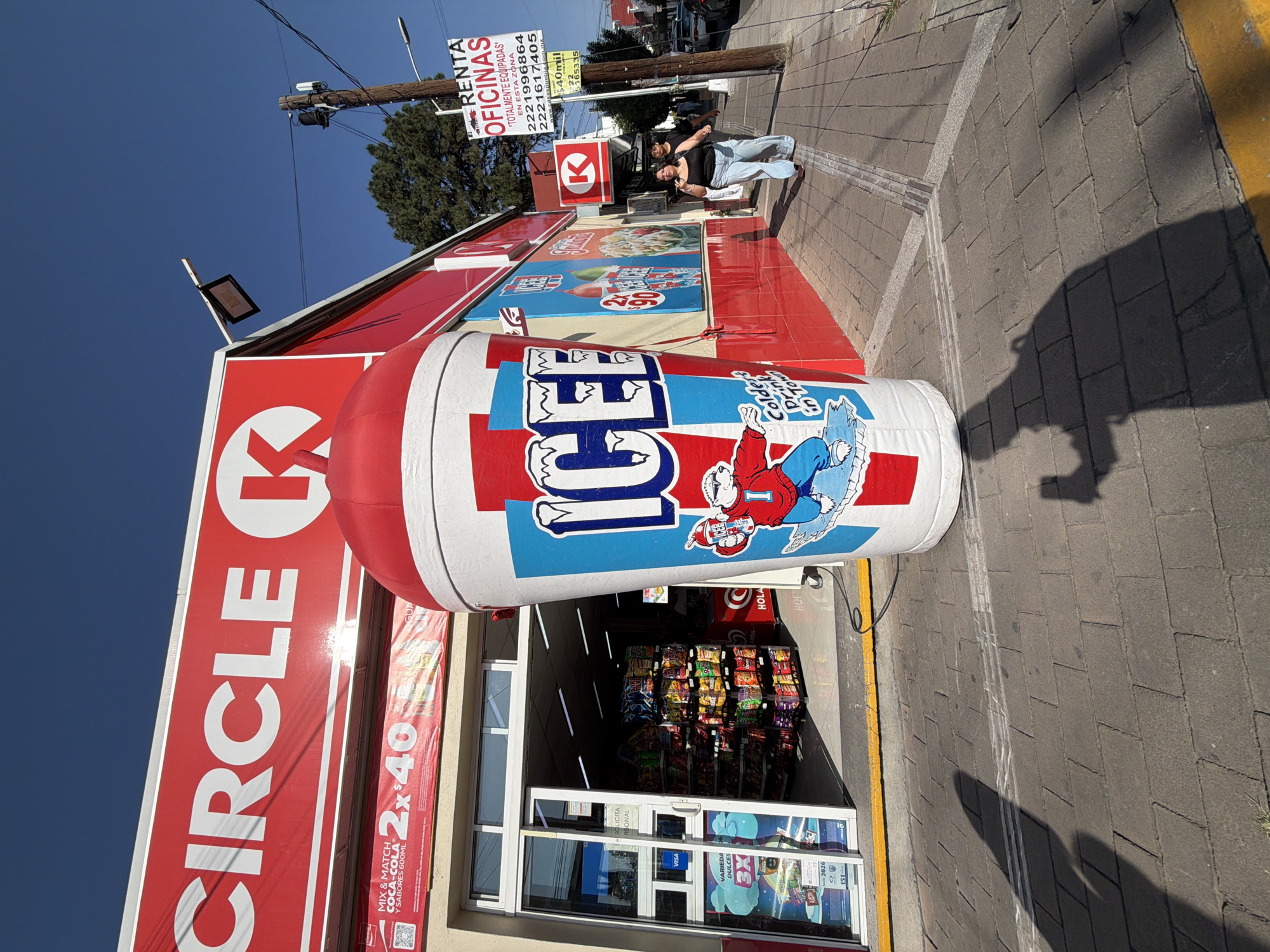A Circle K convenience store is shown in Puebla, Mexico. The building is red and white with the Circle K logo prominently displayed. An inflatable ICEE cup stands in front of the store. Two individuals are standing on the sidewalk next to the store. A sign advertising office rentals is visible. The environment includes a clear blue sky and typical urban street elements such as sidewalks and utility poles.