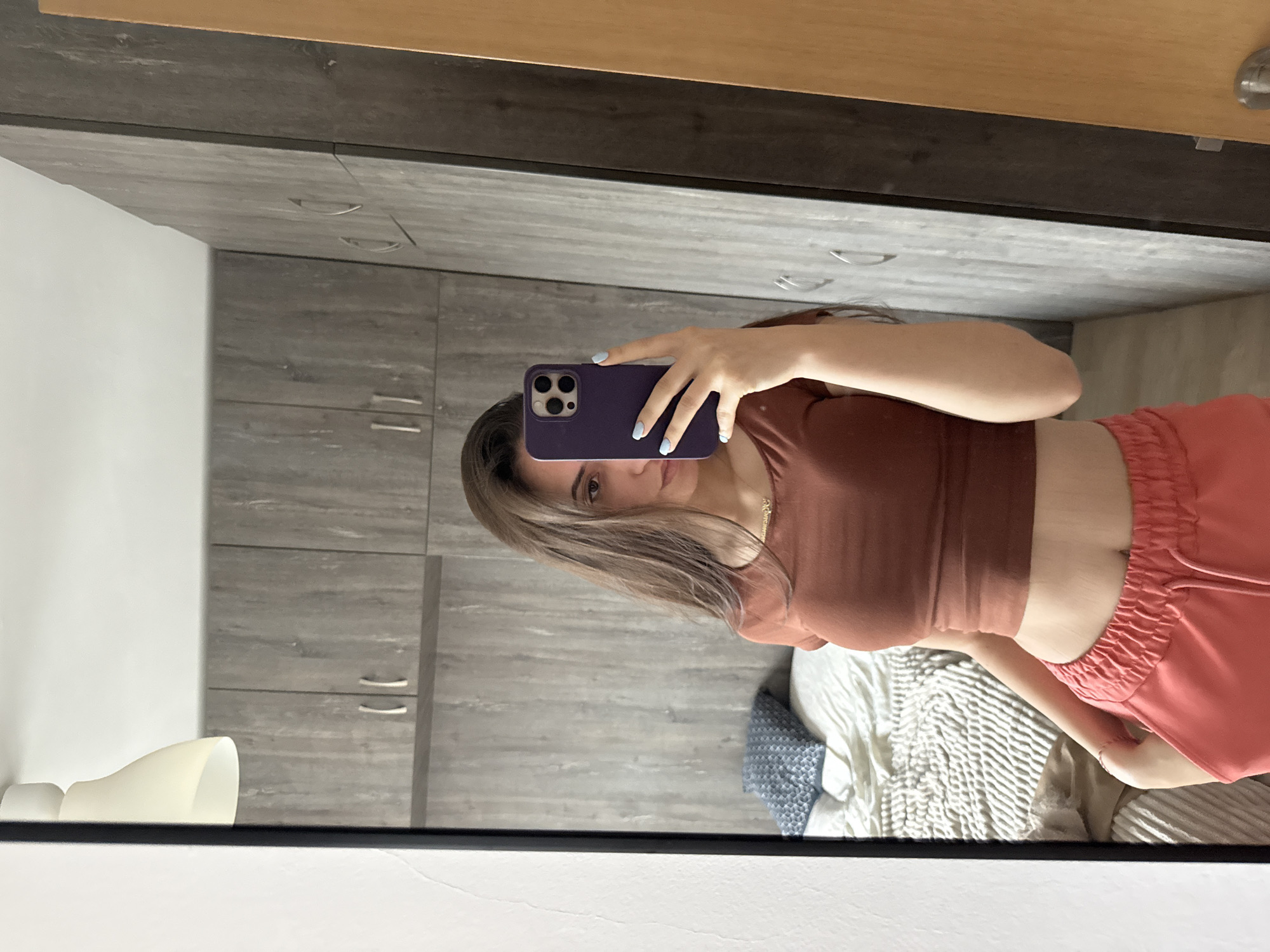 A woman is taking a selfie in a mirror. She is wearing a maroon crop top and salmon-colored shorts. Her hair is light brown. She is holding a purple iPhone in front of her face. The background shows a grey cabinet and a bed with a grey and white duvet. The location cannot be determined.