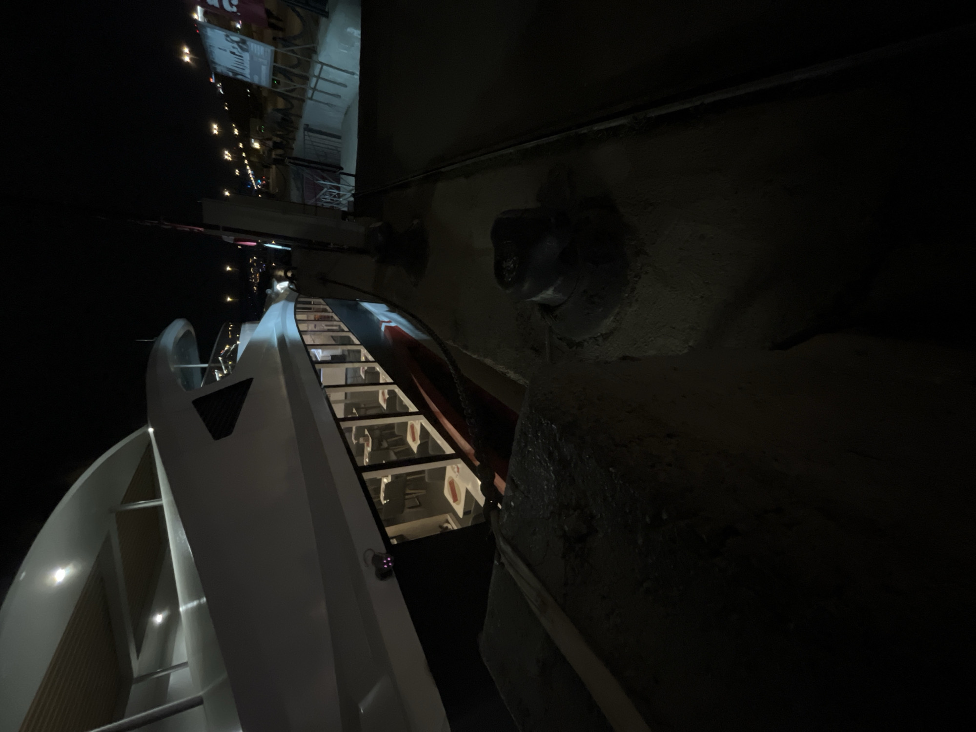 A low-angle, rotated image shows a river cruise boat docked against a cement embankment at night in Unknown, Unknown. The boat has a white exterior with a glass-enclosed cabin featuring rows of illuminated seating visible through the windows. The embankment is dark and textured, with large, dark protrusions or fixtures attached to it. In the background, lights from buildings or structures are visible along the river. The overall lighting is dim, with the main sources of illumination coming from the boat's interior and the distant city lights.