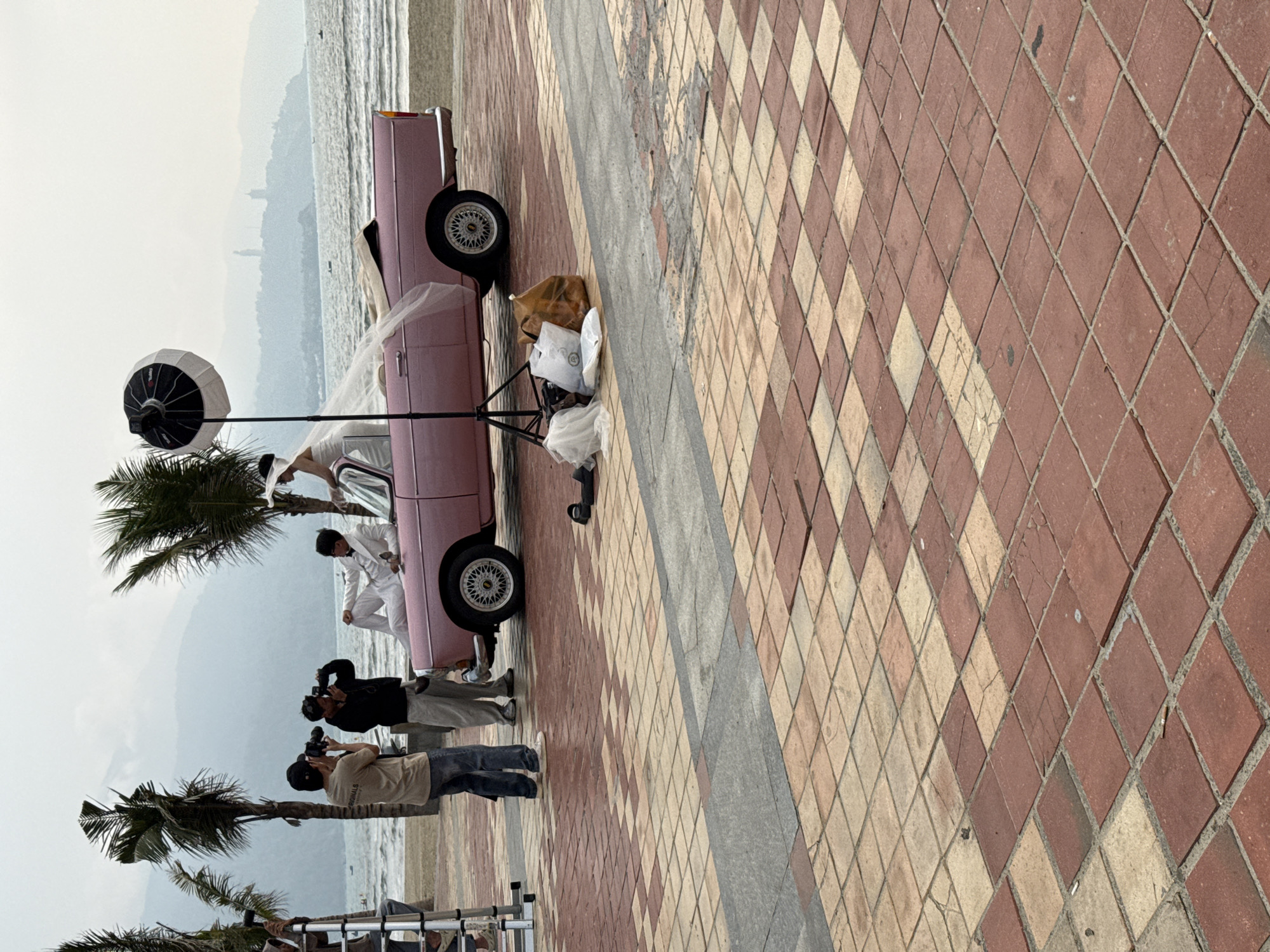 The image depicts a professional photoshoot in progress on a paved promenade bordering a sandy beach in Da Nang, Vietnam. The background features the South China Sea leading to a hazy mountainous coastline, notably identifying the Son Tra Peninsula by the distant white Lady Buddha statue.
A light pink vintage convertible car, with its soft top down, is positioned on the beach side of the promenade. A male subject, attired in a white suit, black bow tie, and sunglasses, reclines on the car's hood. A female subject, wearing a white wedding gown, a long veil draped over the car's rear, and a wide-brimmed white hat, is leaning into the driver's side of the vehicle, her back partially to the viewer.
Two male photographers are actively capturing the scene. One, in a beige t-shirt and jeans, holds a camera centrally. The second, in a dark hooded top, stands to his right, also operating a camera. A third individual is partially visible on a silver ladder to the far left, suggesting additional crew. Photography equipment includes a studio strobe light with a white parabolic softbox reflector on a stand, situated to the right of the car. Bags and discarded white fabric (possibly part of the veil) are on the ground near the light.
The promenade surface consists of a checkered pattern of reddish-brown and beige square tiles, separated by a strip of grey rectangular tiles. The tiles show signs of wear, cracking, and dampness. Several tall palm trees line the beach behind the photoshoot, and small boats are visible on the water. The sky is overcast or hazy.