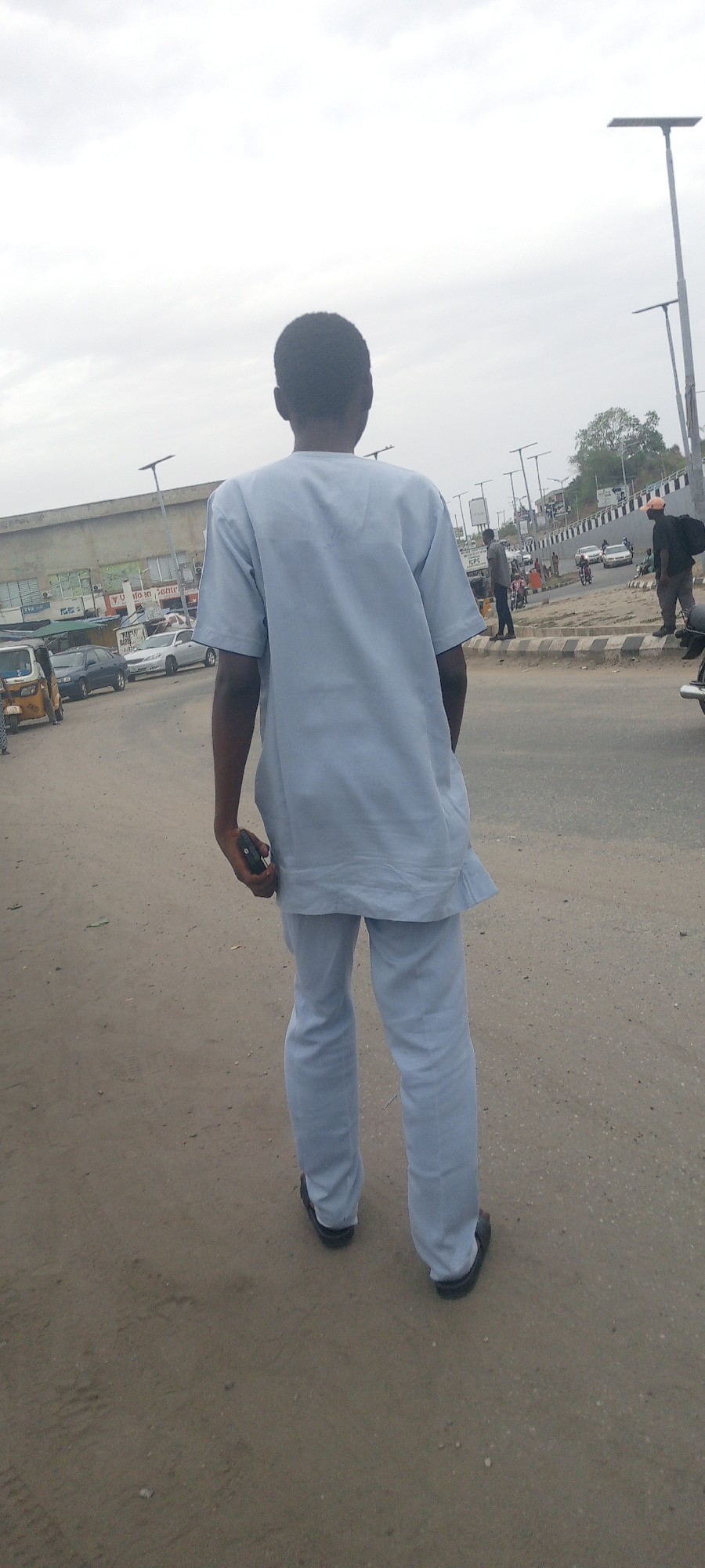A man in light blue clothing stands on a dirt and paved road in Adankolo, Nigeria, facing away from the camera. He holds a dark object in his right hand. In the background, there is vehicular traffic, including cars, a motorcycle, and an auto-rickshaw. There are other individuals visible on the sidewalk near a multi-story building with signage. Street lights are present along the road, and the sky is overcast.