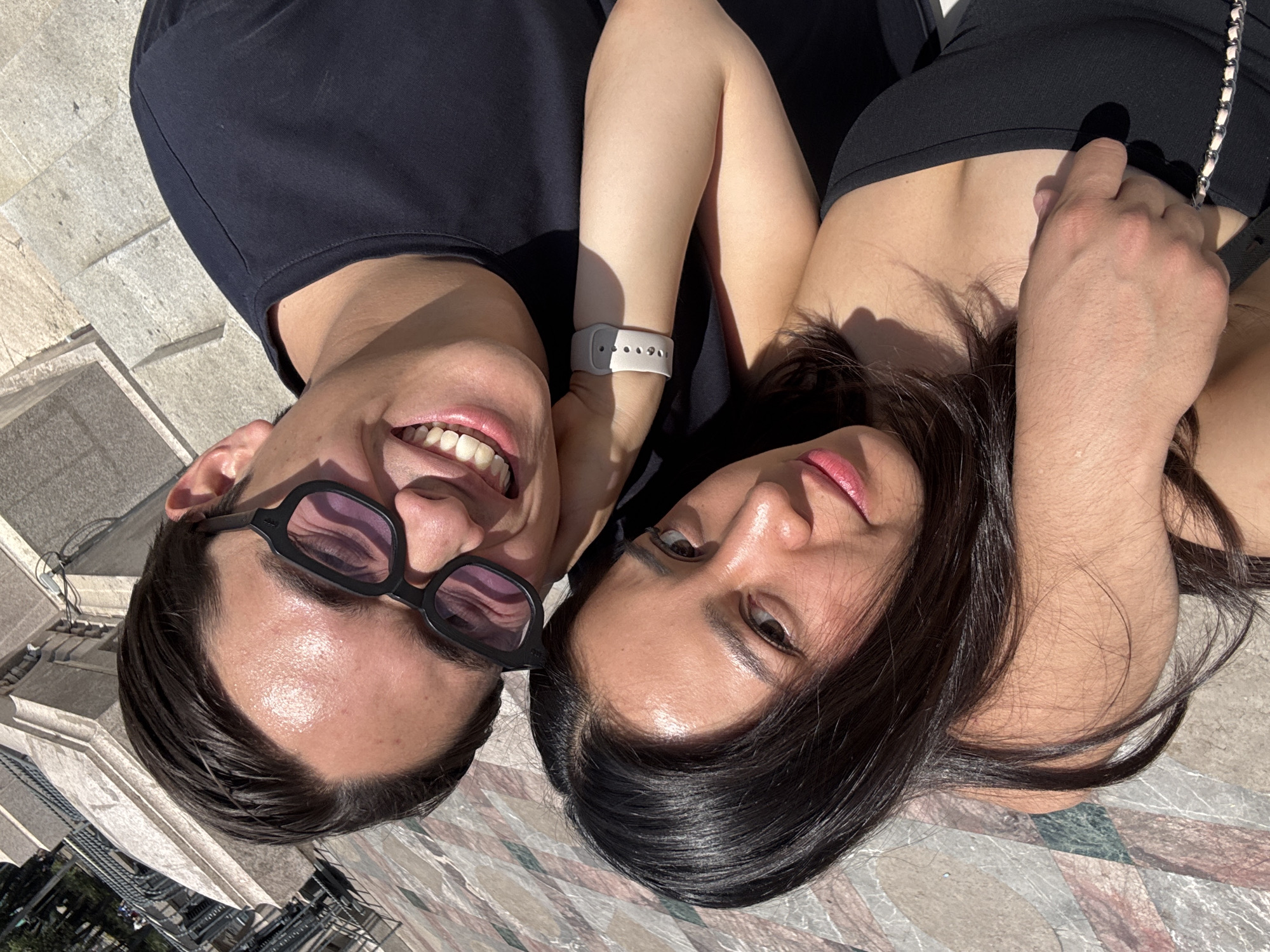 A medium-shot, eye-level photograph shows a couple lying on the ground, taking a selfie. The man is on the left with dark hair and dark-framed sunglasses, smiling broadly and showing his teeth. He is wearing a dark-colored shirt. The woman is on the right, with dark hair and a fair complexion. She is looking at the camera with a slight smile. Her arm is visible, adorned with a white smartwatch. Both individuals are lying close together. The location seems to be a public area, as suggested by the patterned stone ground. Based on the distinctive architecture visible in the blurry background, including building structures, the scene may be in Mexico City, Mexico.