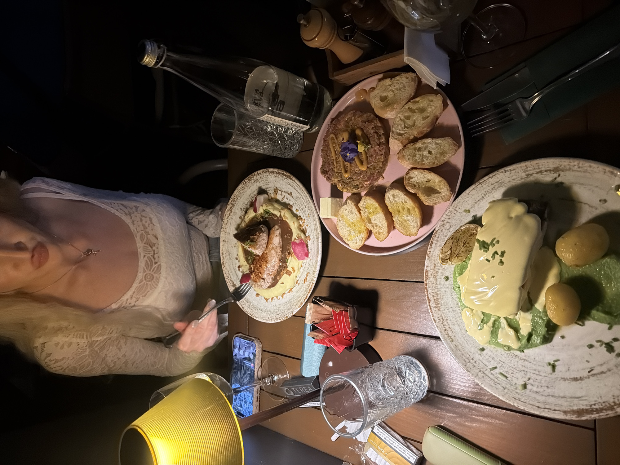 The image depicts a person in a white lace dress dining at a table with multiple dishes in what appears to be a restaurant, possibly in Belgrade, Serbia. The table holds three plates: one with beef tartare and bread, another with a meat dish featuring mashed potatoes and reddish garnishes, and the third with what appears to be eggs benedict with potatoes and greens. A bottle of water, glasses, cutlery wrapped in green napkins, a salt shaker, and various small items like sauce packets and cigarette packaging are also on the table. A smartphone leans against a glass, possibly being used for lighting or entertainment. The lighting is dim with a warm-toned lamp providing illumination.