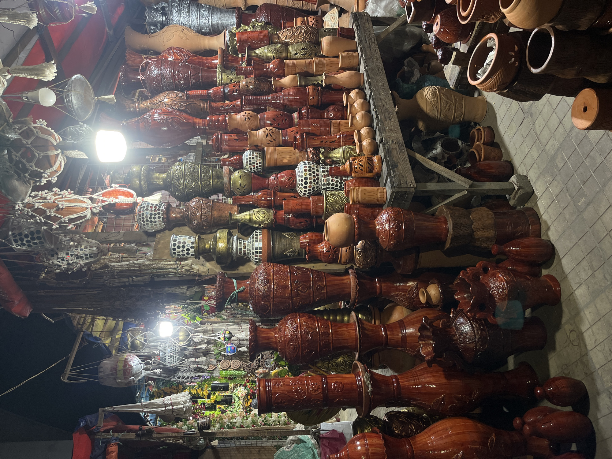The image depicts a crowded marketplace stall in Dhaka, Bangladesh, filled with an array of ceramic and clay pots, vases, and decorative items. The items are arranged in rows, with various shapes, sizes, and colors, ranging from earthy brown tones to ornate metallic and mosaic designs. The stall is illuminated by overhead lighting, which casts light and shadows across the merchandise. There are also other items such as hanging light fixtures. The backdrop hints at the bustling environment of a marketplace with people and other stalls present.
