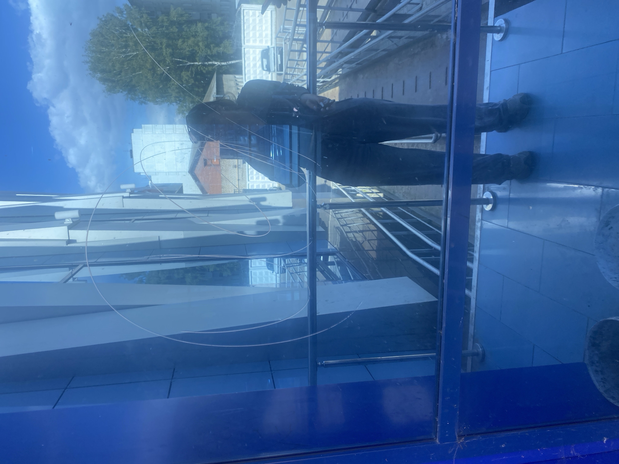 A person, appearing to be of dark complexion with dark hair, stands inside a building, reflected in a blue-tinted glass window. They are wearing dark clothing and dark shoes. The window has visible scratches and streaks. Outside the window, a walkway with metal railings is visible. Beyond the walkway, there are buildings, trees, and a blue sky with clouds. The scene appears to be outdoors, possibly in an urban or semi-urban environment. The location is identified as Chaikovsky, Russia.