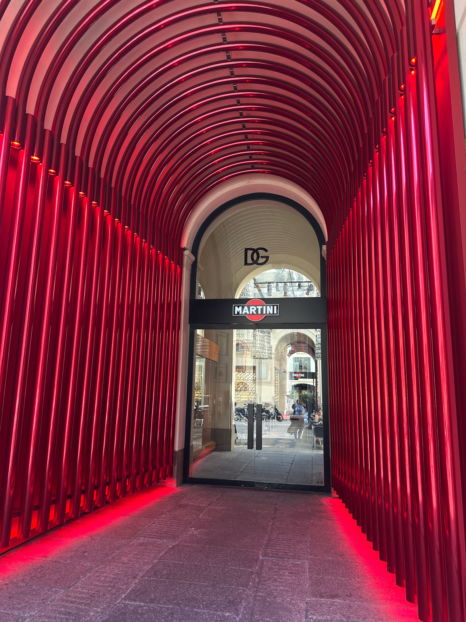 The image shows an arched walkway with a vibrant red, ribbed metal structure lining both sides and the ceiling. The walkway leads to an entrance with glass doors, above which are two signs: one with "DG" logo, and below it, a sign that reads "MARTINI" with its circular red logo. Through the glass doors, an interior space is visible, with what appears to be a bar or lounge area. Beyond that, another archway reveals a glimpse of a street scene with motorcycles parked and people walking. The floor of the walkway is paved with stone tiles, and a red light emanates from the base of the metal structure onto the ground. The architectural style suggests a location in Milan, Italy, likely a commercial establishment or a public passage designed with a striking aesthetic.