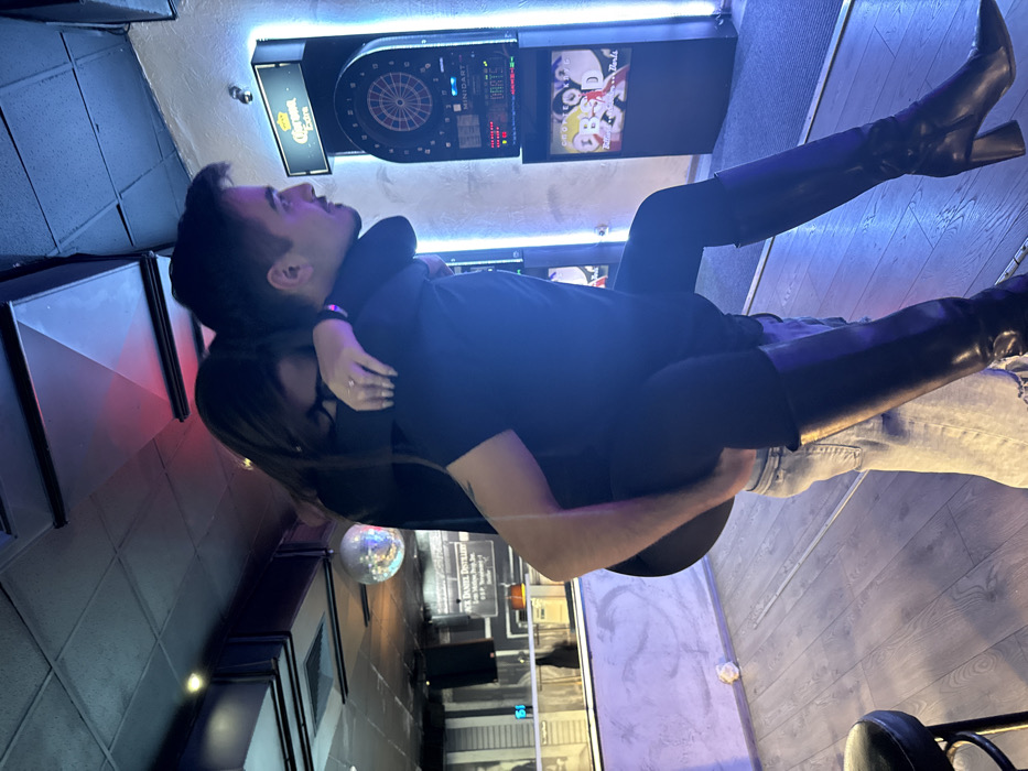 The image depicts an indoor scene within an entertainment venue, likely a bar or pub, in Sofia, Bulgaria. Two individuals are present: a male carrying a female on his back in a piggyback ride.
The male, positioned on the right and facing towards the right in profile, has dark hair, wears a dark short-sleeved top and light-wash jeans. His right arm is wrapped around the female's legs for support, while his left arm is positioned around her upper thighs. The female, situated on the male's back, has long dark hair partially obscuring her face, wears a dark long-sleeved top, dark leggings, and knee-high black boots with block heels. A ring is visible on her left ring finger. Her arms are wrapped around the male's neck and shoulders, and her legs are wrapped around his waist, with her head resting against his upper back.
The environment includes a light-colored wooden floor and a dark, possibly tiled, ceiling featuring recessed lighting and visible air ducts. A disco ball hangs from the ceiling in the left background. A black upholstered stool is visible in the bottom left foreground. In the left background, a bar area features a light-colored counter and a wall-mounted sign displaying "Jack Daniel's Distillery." In the right background, an electronic dartboard machine, labeled "MINIDART," is present, framed by vertical blue LED lighting strips. Above the dartboard, an illuminated "Corona Extra" sign is mounted. A screen on the dartboard displays text including "DARTS," "MINIDART," and "OBSD." The overall lighting is dim, with prominent blue and red accent LED lights contributing to the atmosphere, while the dartboard and bar area are more brightly illuminated.