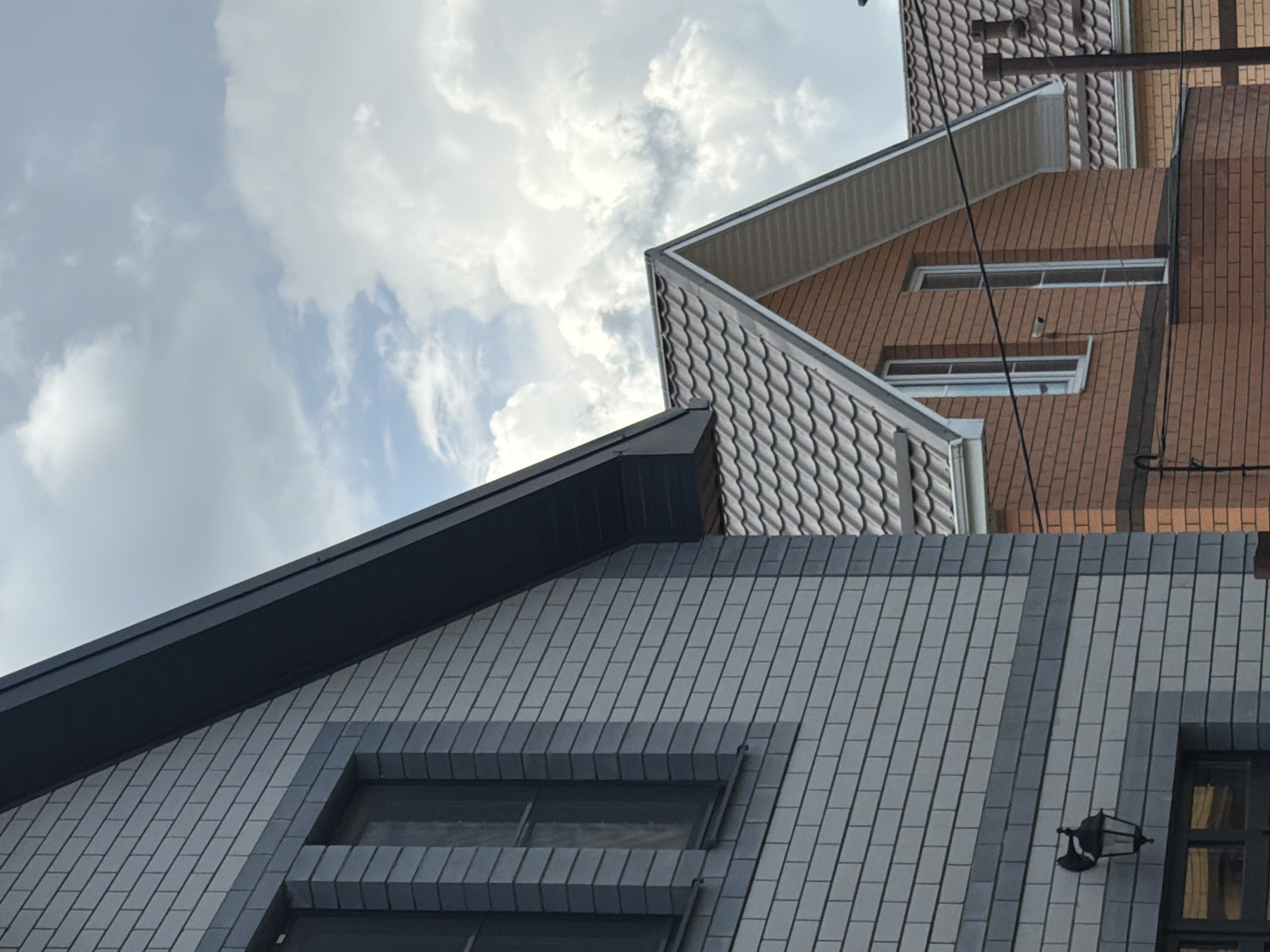 The image shows a low-angle view of two adjacent buildings against a cloudy sky. The building in the foreground has a gray brick facade and a dark gray roof. Two dark-framed windows are visible, along with an exterior light fixture mounted on the wall. The second building, partially visible behind the first, has a brown brick facade and a gray metal tile roof. Two windows are visible on this building. The location is Rossiyskiy, Russia.