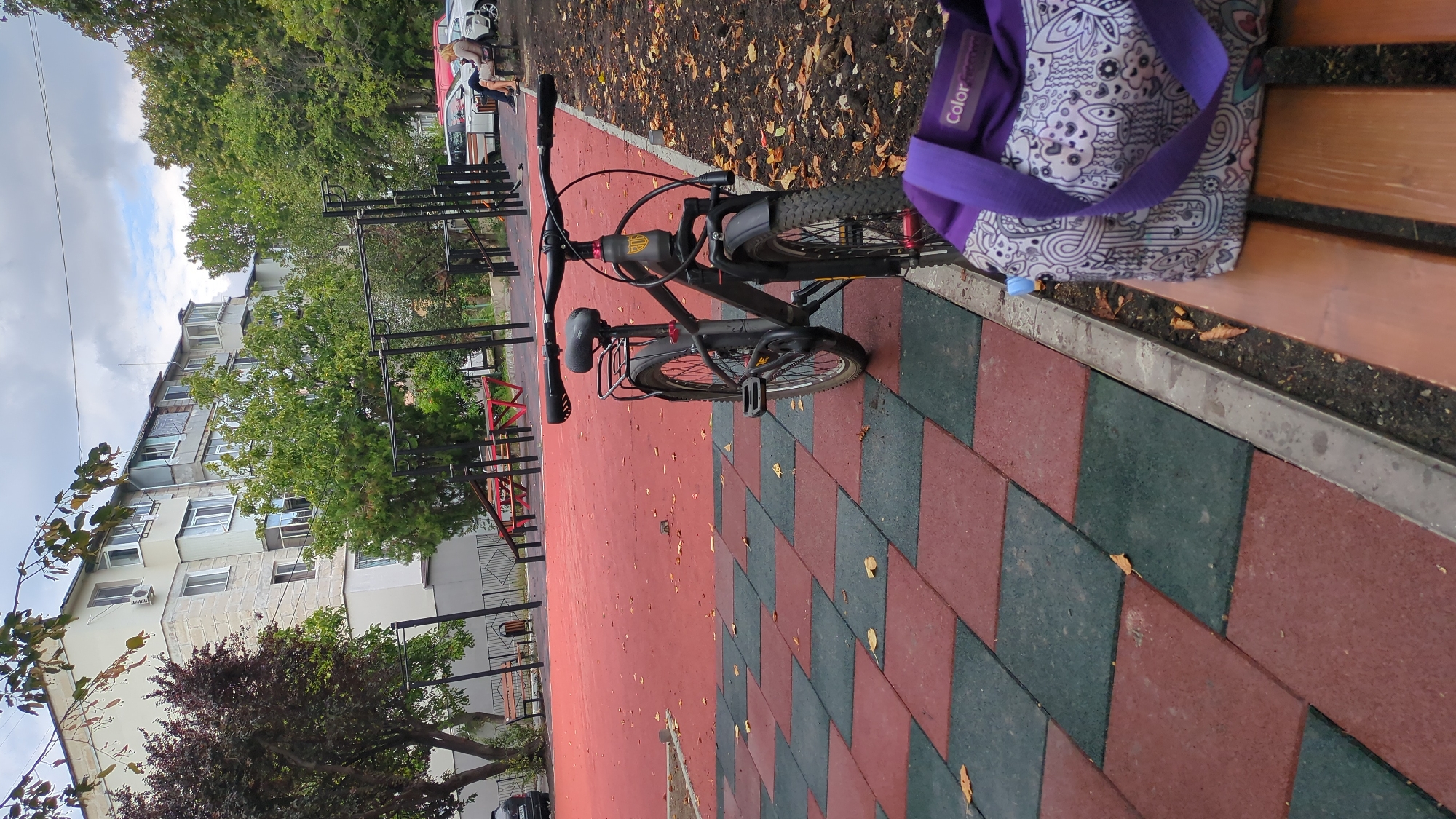 The image depicts an outdoor scene in Chișinău, Moldova, likely a public park or recreational area. In the foreground, a dark-colored bicycle stands on a paved area with red and green interlocking rubber tiles. To the right, a wooden bench is visible, and on it rests a patterned bag with purple straps. Behind the bench, dry leaves are scattered on the ground. The middle ground features a red-colored sports surface, a metal workout structure with horizontal bars, and some red benches and tables. In the background, a multi-story apartment building with light-colored facades and balconies is situated amidst lush green trees. The sky is overcast with clouds. A few people are seated on a bench near cars in the distance. The overall impression is of an urban park setting during autumn.