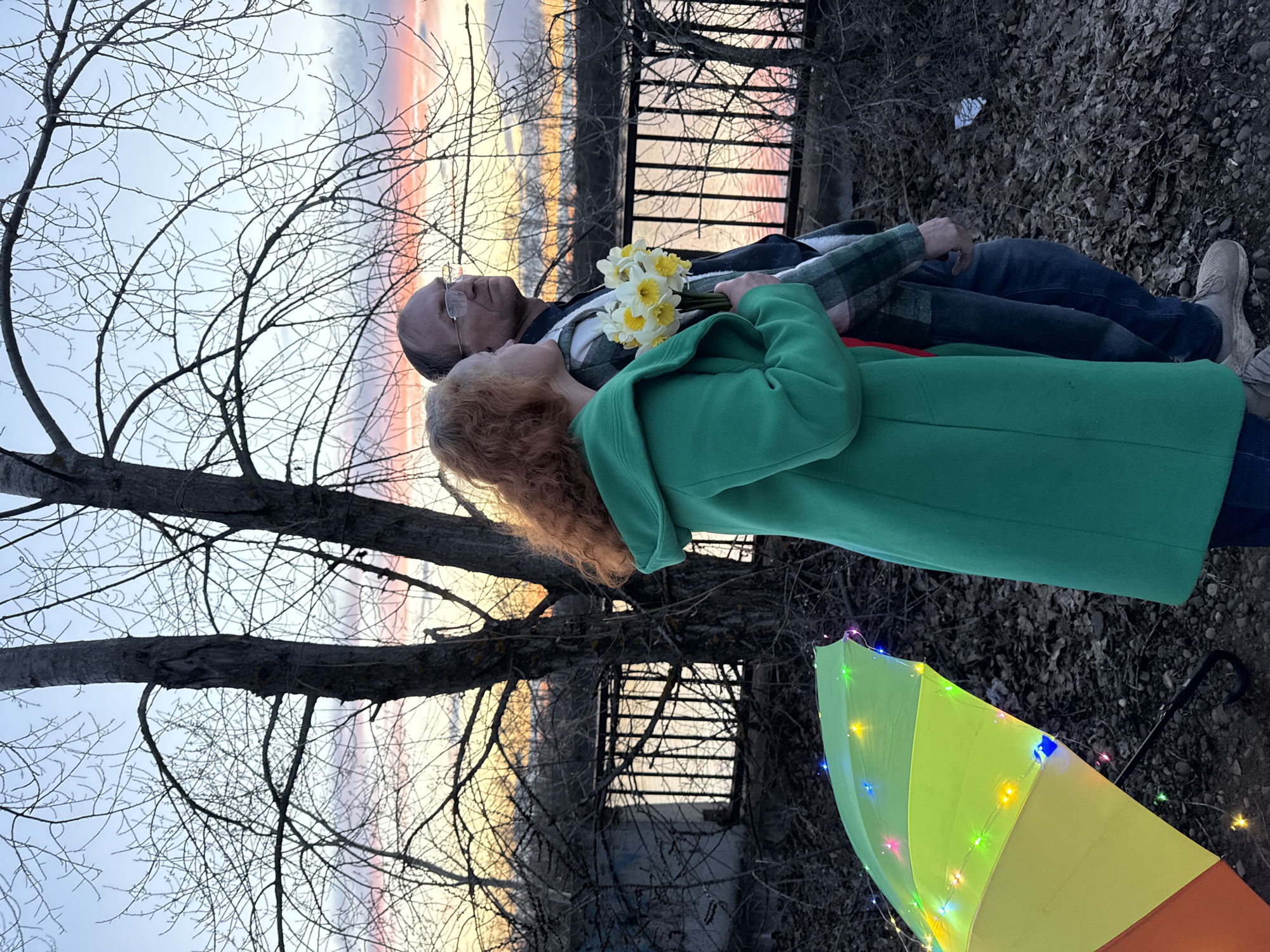 An eye-level outdoor photograph captures an intimate moment between two individuals at sunset or sunrise in Chaikovsky, Russia. An older man and a woman stand close together, looking towards a vibrant sky painted with hues of pink, orange, yellow, and blue. The man, wearing glasses, a plaid shirt, and dark jeans, has his right arm wrapped around the woman's back. The woman, with reddish-blonde curly hair, is dressed in a bright green coat and holds a bouquet of yellow and white daffodils to her chest.
They stand on ground covered with dry, fallen leaves, suggesting late autumn or early spring. Behind them, a dark metal fence or railing runs horizontally, possibly overlooking a body of water that reflects the colorful sky. Bare tree branches, intricately silhouetted against the bright sky, frame the scene, dominating the upper left and also appearing in the mid-ground. To the lower right, a rainbow-colored umbrella, adorned with small, multi-colored LED lights, lies open on the ground. The overall mood is peaceful and contemplative, capturing a serene moment between the couple amidst a beautiful natural backdrop. No visible text is present.