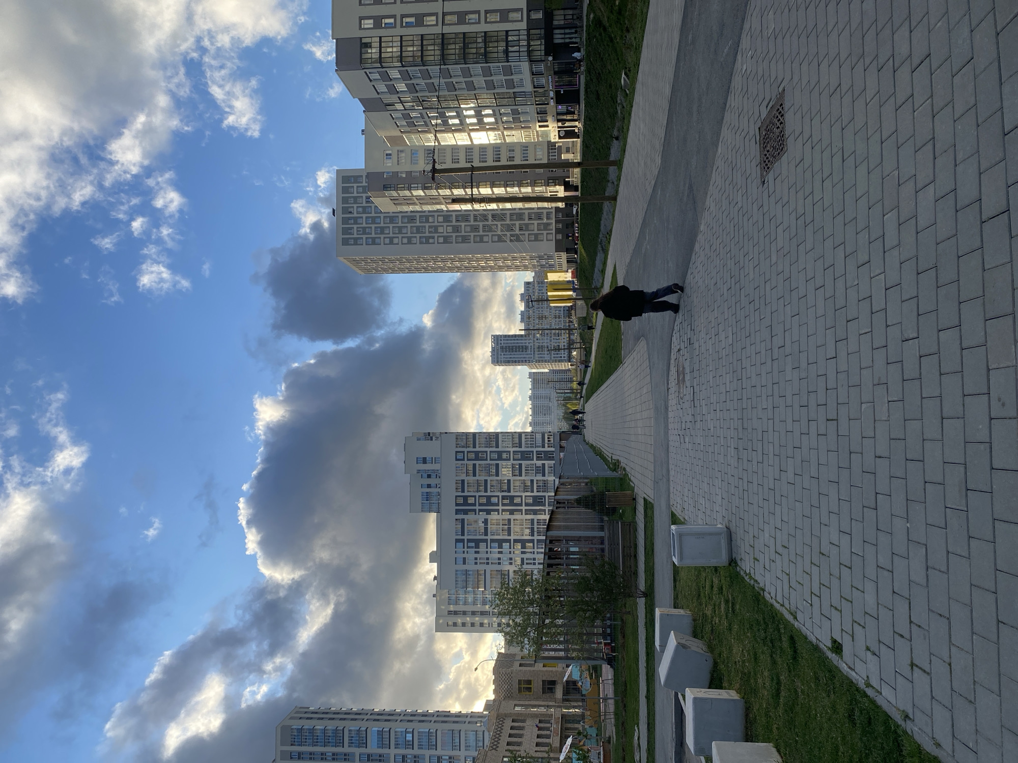 A streetscape in Yekaterinburg, Russia, reveals a modern urban environment. Multiple high-rise apartment buildings line the left side of the frame, reflecting sunlight. A cloudy sky occupies the upper portion of the image. A person is seen on a paved walkway, and landscaping featuring grass and concrete blocks runs parallel.