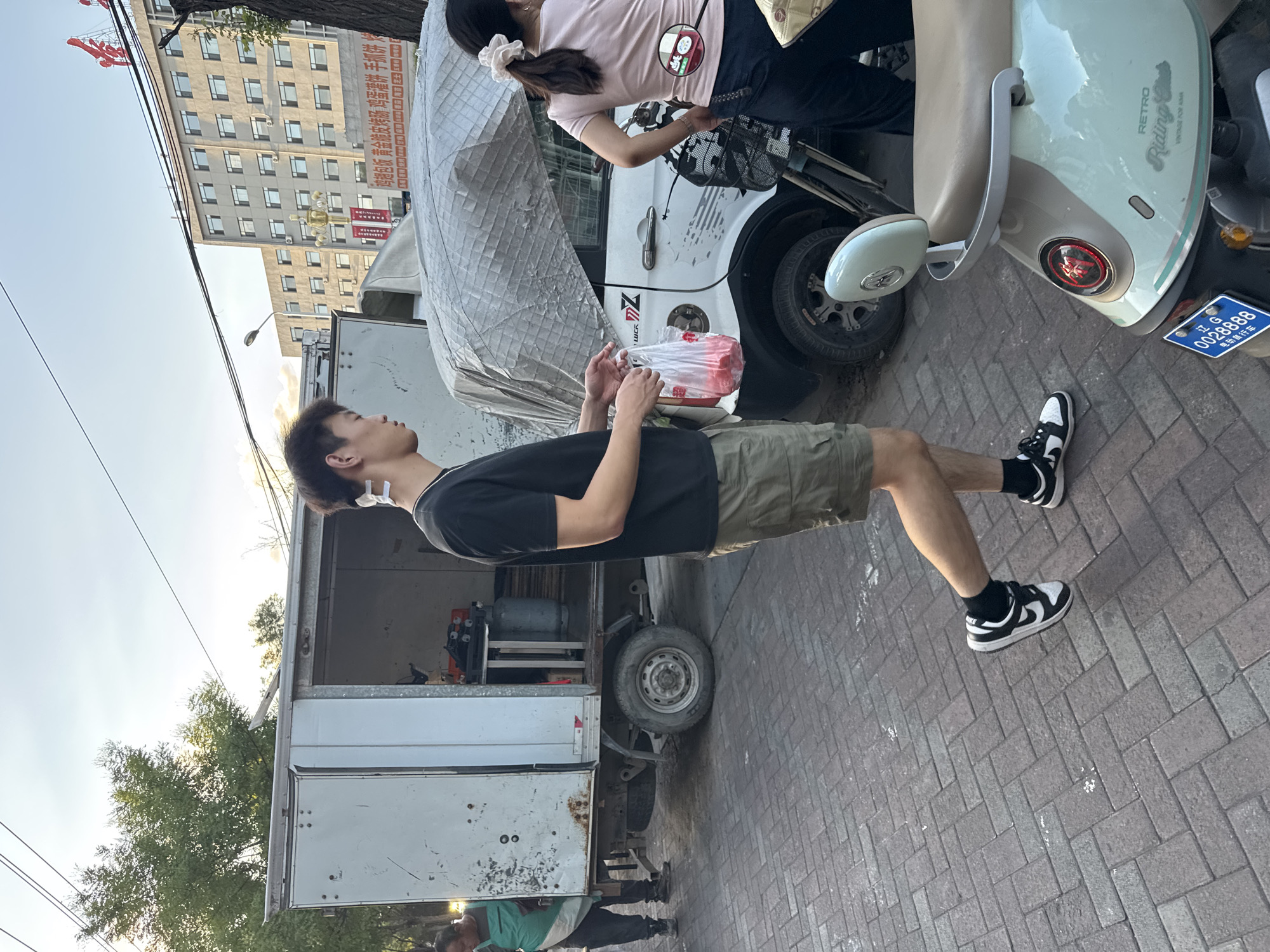 In Zhongtun, China, on a paved street, a young man in a black t-shirt and khaki shorts walks past a delivery truck and a white van covered with a tarp. He is holding a plastic bag filled with red items. To the right, a woman with her hair in a ponytail, wearing a pink shirt and dark pants, stands near a scooter. The background shows a multi-story building with many windows and some trees. The sky is clear.