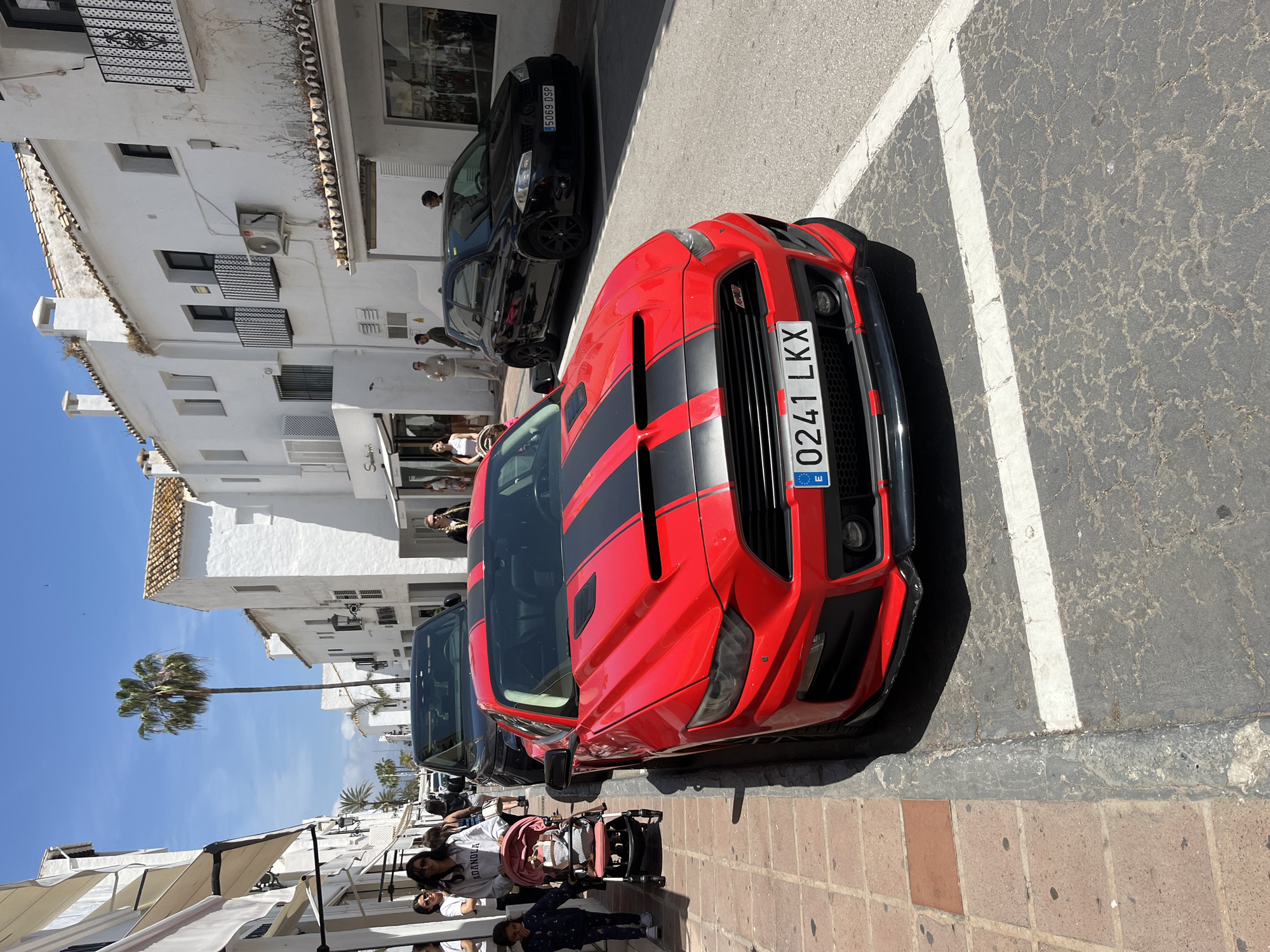 The image depicts a street scene in Nueva Andalucía, Spain, with a red Ford Mustang with black racing stripes parked on the right side of the road. The car's license plate reads "0241 LKX". A black car is parked behind the Mustang. The road is marked with white parking lines. On the left side of the road, there are white buildings with multiple levels, balconies, and air conditioning units. People are walking on the sidewalk, including a person pushing a stroller. A palm tree is visible in the background. Awnings extend from buildings on the left. The weather appears sunny.