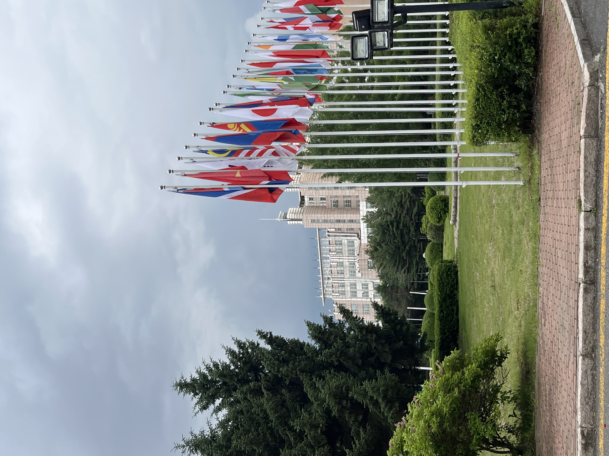 The image captures a view of a row of international flags displayed on white poles in front of a large, multi-story building. The building appears to be modern, with many windows and visible piping on its exterior. Tall evergreen trees are prominent in the foreground, with well-maintained green lawns and hedges surrounding the flagpoles. A brick pathway runs along the bottom of the image. The sky is overcast with gray clouds. This scene is located at the entrance of the Jilin Provincial Science and Technology Museum in Changchun City, China.