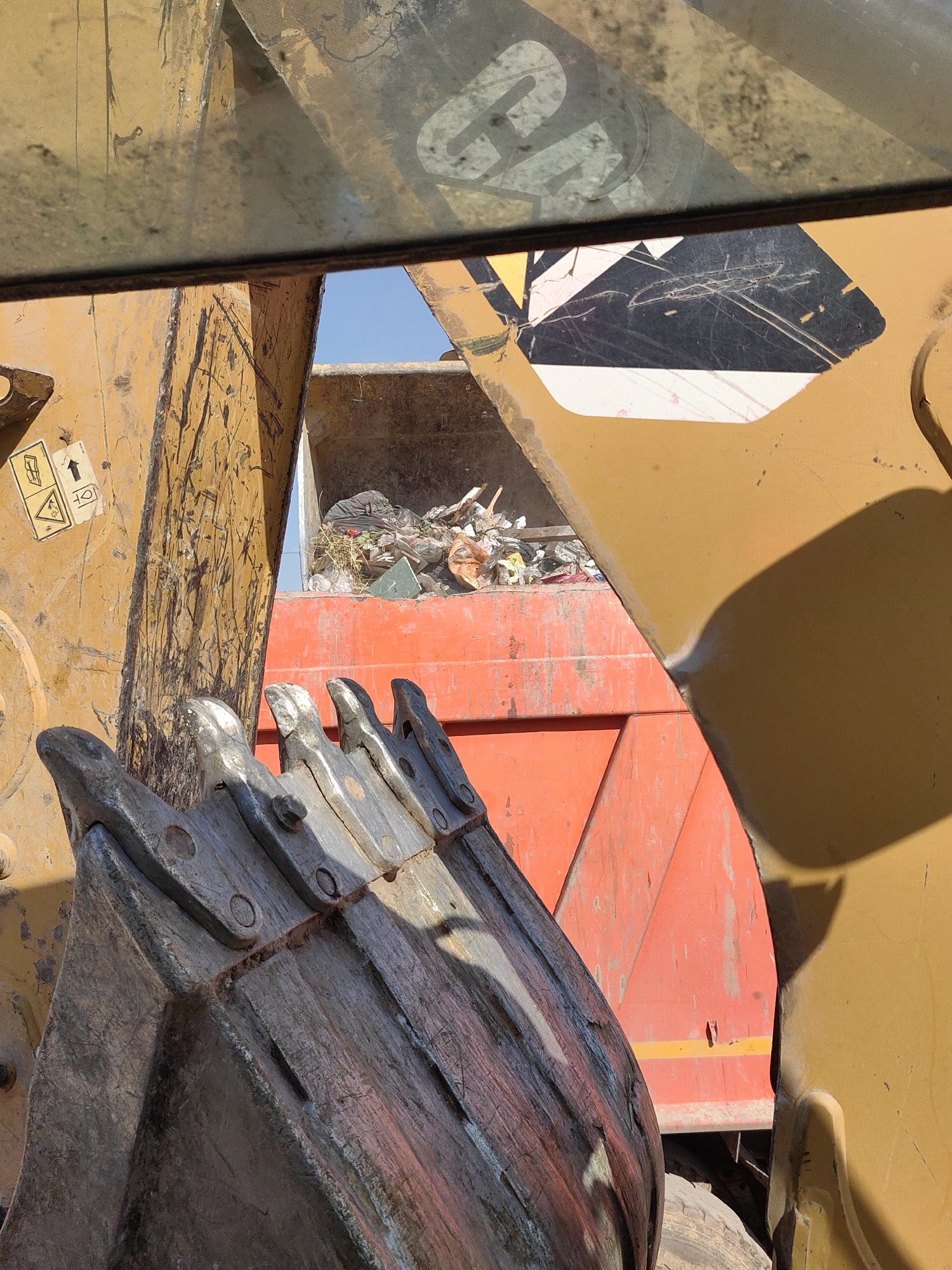 An extreme close-up view from the perspective of an excavator operator shows a pile of trash in the bed of a dump truck. The excavator's bucket, with its teeth visible, is in the foreground, partially obscuring the view. The dump truck is orange, and the trash includes various plastic bags and debris. The scene appears to be outdoors under a clear blue sky, suggesting it could be a landfill or a waste disposal site. The location is stated as Shymkent, Kazakhstan.